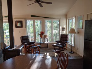 A spacious living room with large windows, a ceiling fan, and a dining area with a table and chairs. - Treehouse on the river. (Au Train)