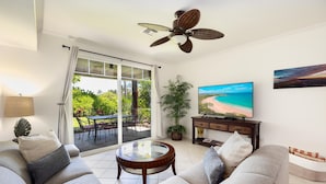 Ein Wohnzimmer mit einer Couch, einem Glastisch, einem Fernseher und Blick auf einen Strand durch eine Schiebetür.