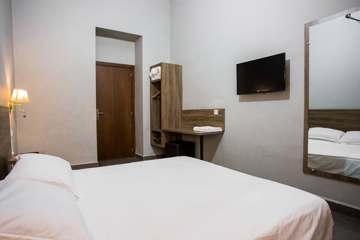 A hotel room with a bed, a TV mounted on the wall, a mirror, and a wooden wardrobe.