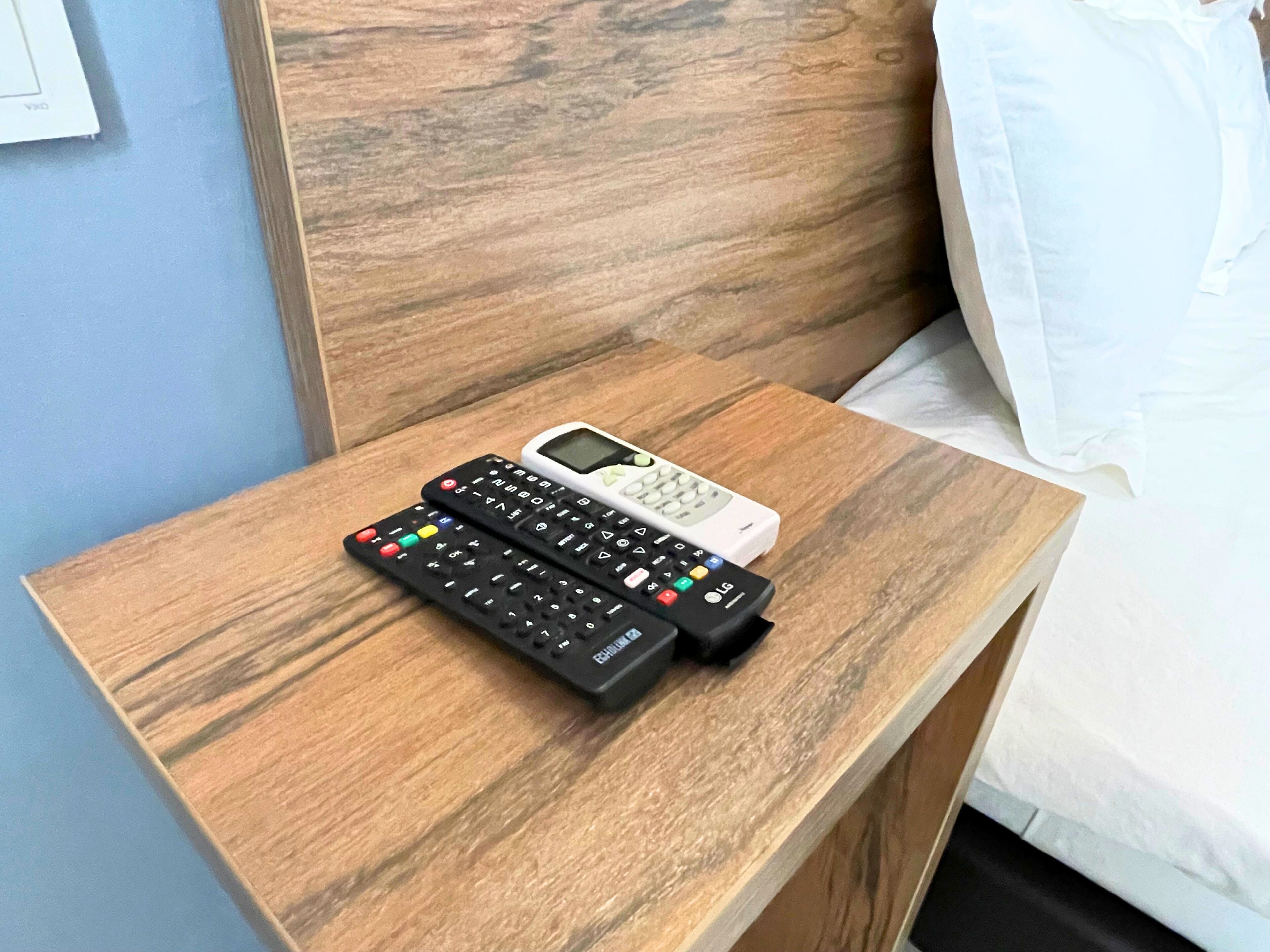 A wooden bedside table with two remote controls and a white device, likely a phone, in a hotel room.