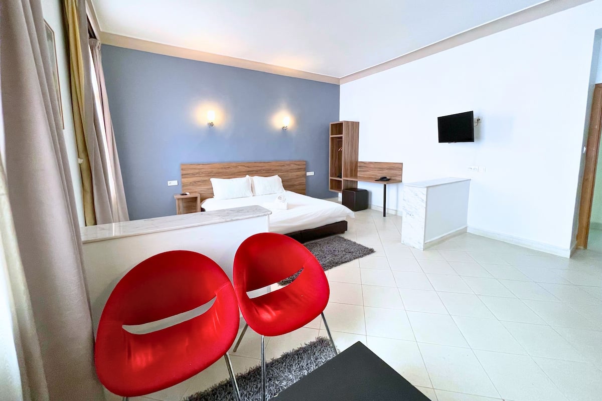 A modern hotel room with a bed, bedside tables, a desk, a TV, and two red chairs.