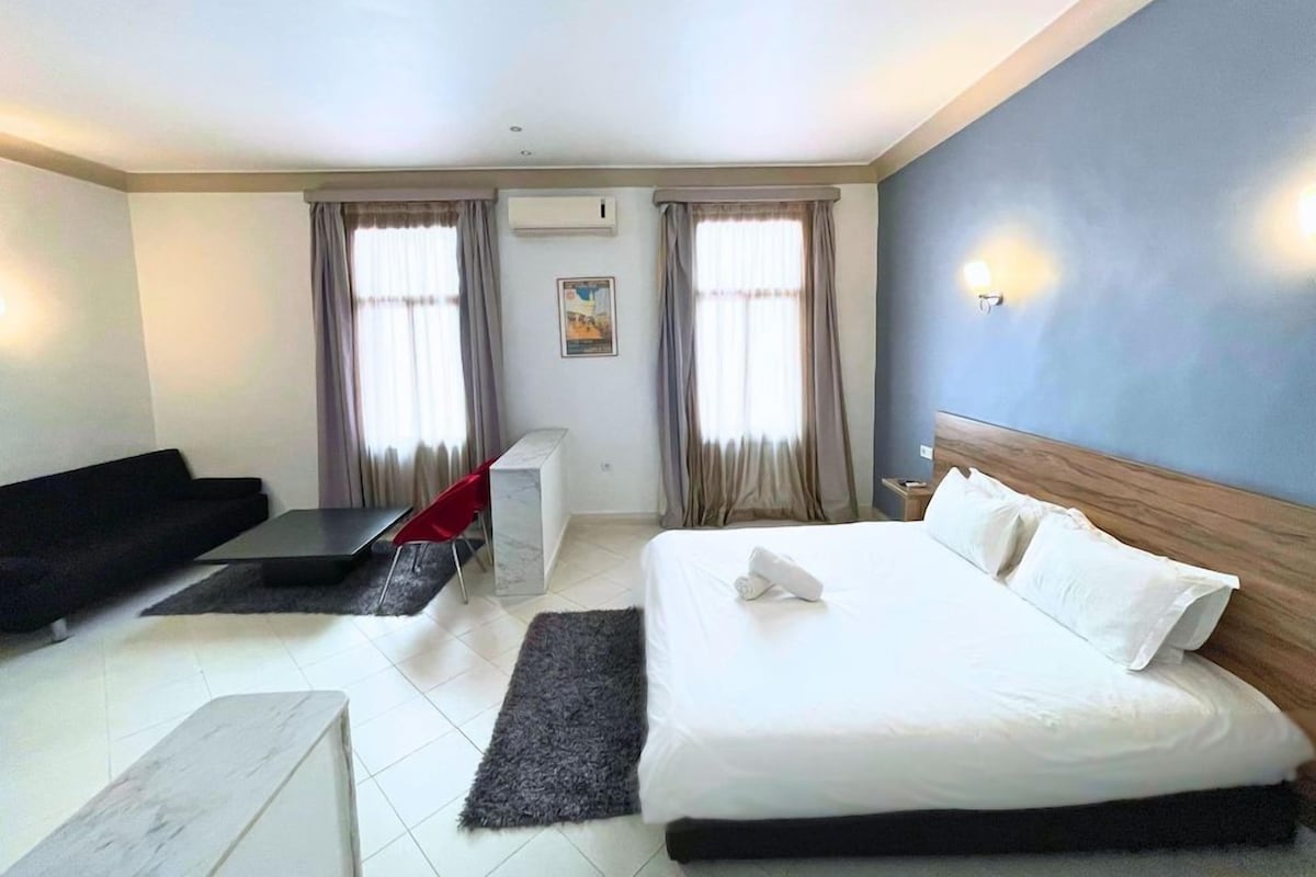 A hotel room with a bed, a sofa, a small table, and a window with curtains.