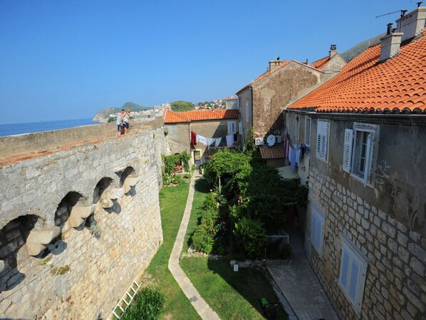 A stone fortification with arched passageways, adjacent residential buildings with red-tiled roofs, and a clear view of the sea.