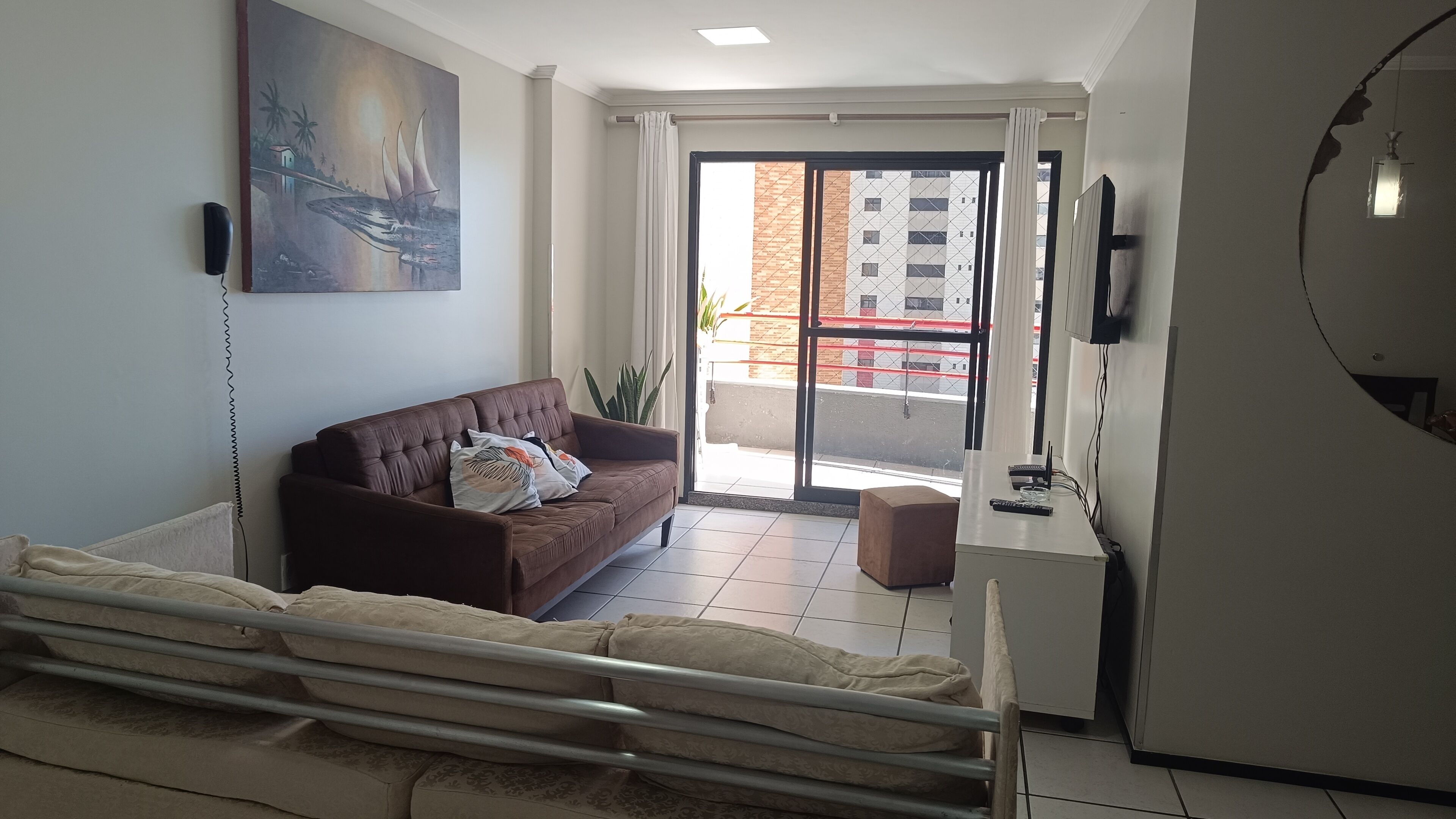 Ein modernes Wohnzimmer mit einer braunen Couch, einem kleinen Tisch, einem Fernseher und einem Balkon mit Blick auf Gebäude.