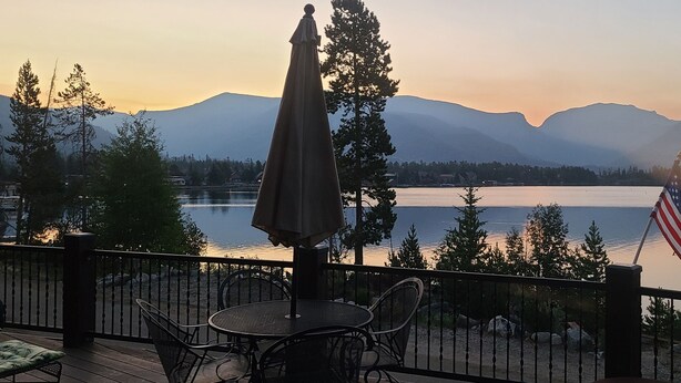 A deck with a table and chairs, overlooking a lake with mountains in the background. - Stunning Lake Views with private boathouse deck, hot tub and close walk to town (Grand Lake)