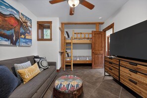 A room with a bunk bed, a television, a sofa with cushions, a small stool, and a ceiling fan.
