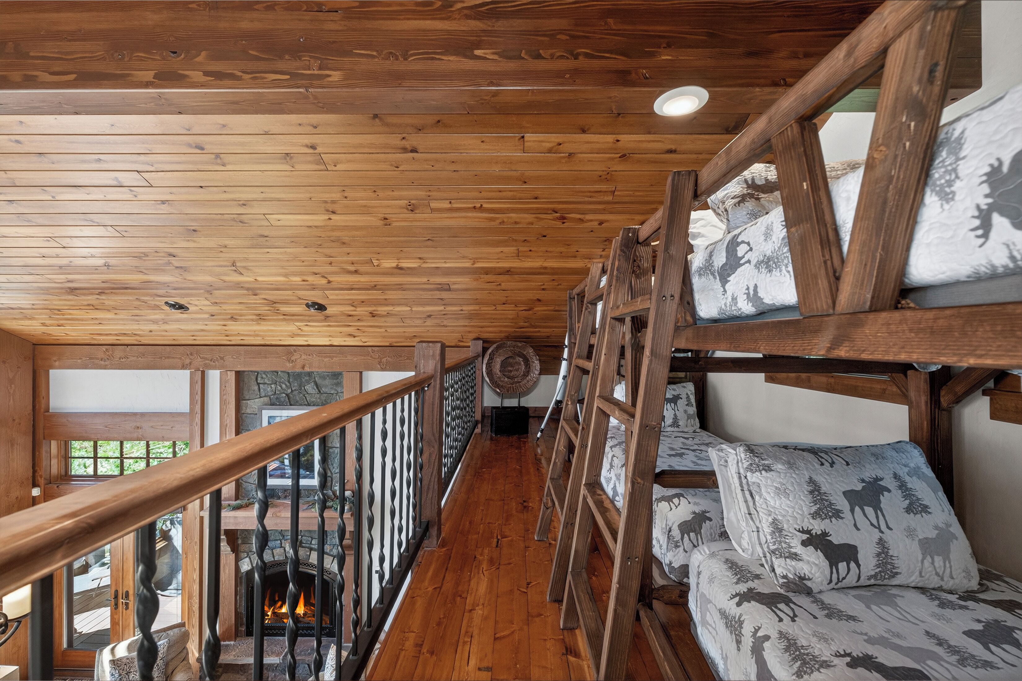 A cabin interior with a wooden staircase, bunk beds with moose-patterned bedding, a fireplace, and a stone wall.