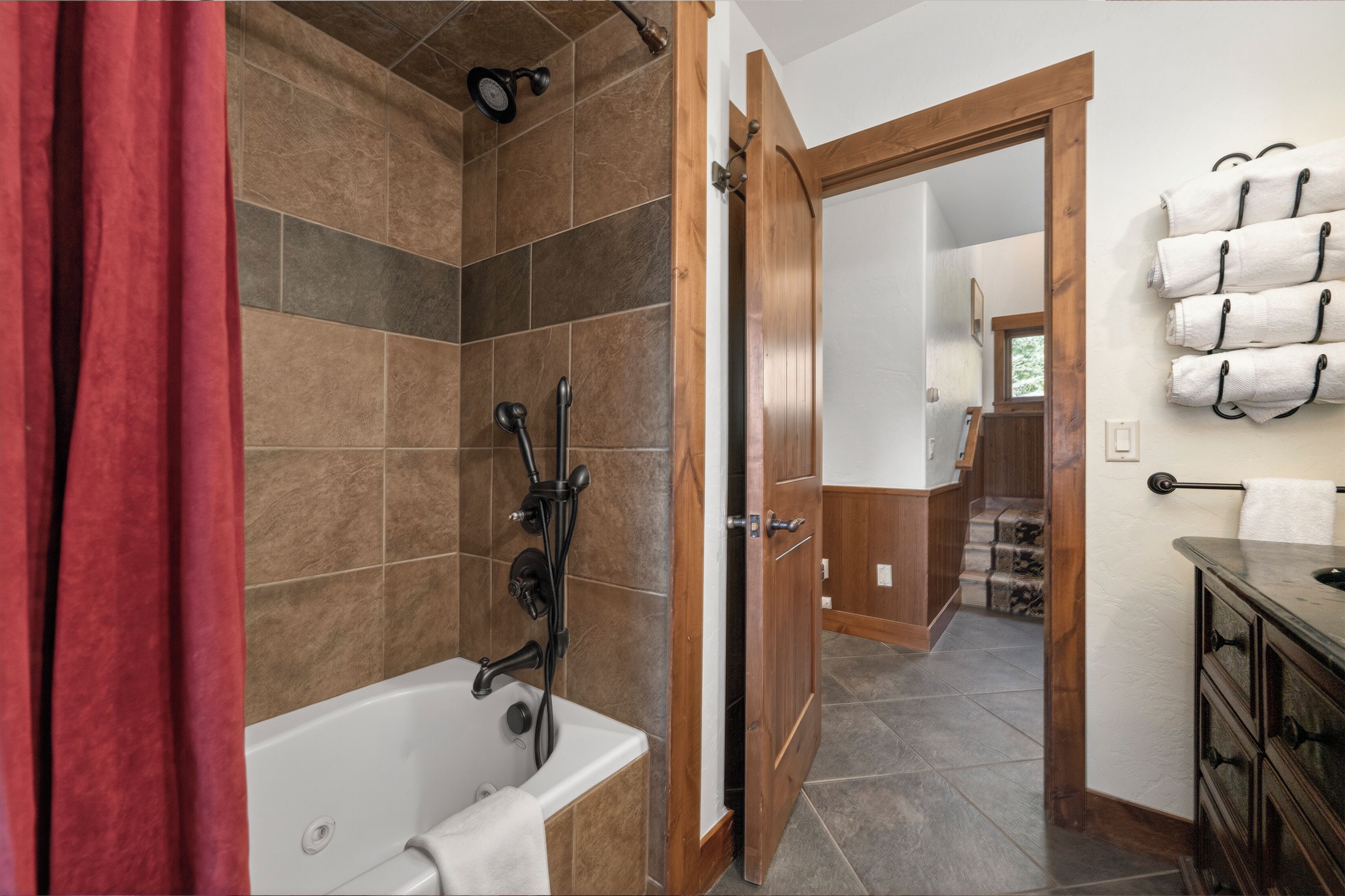 A bathroom with a bathtub, shower, and a wooden door leading to a hallway with a staircase.
