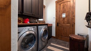 A laundry room with two front-loading washing machines, wooden cabinets, a window with blinds, and a door with a small window. - Stunning Lake Views with private boathouse deck, hot tub and close walk to town (Grand Lake)