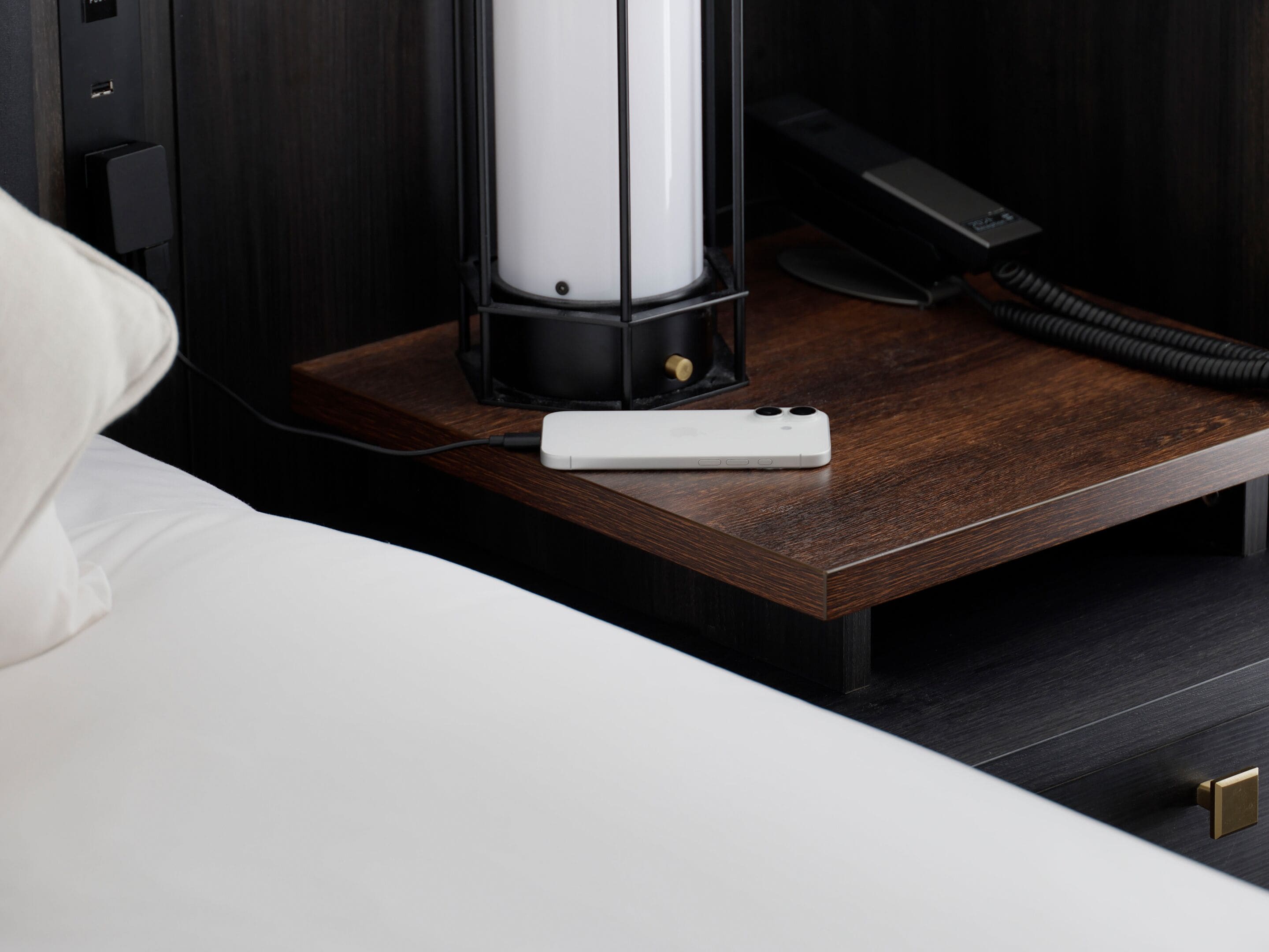 A neatly made bed with a white pillow, a bedside table with a white phone, a black lamp, and a black telephone.