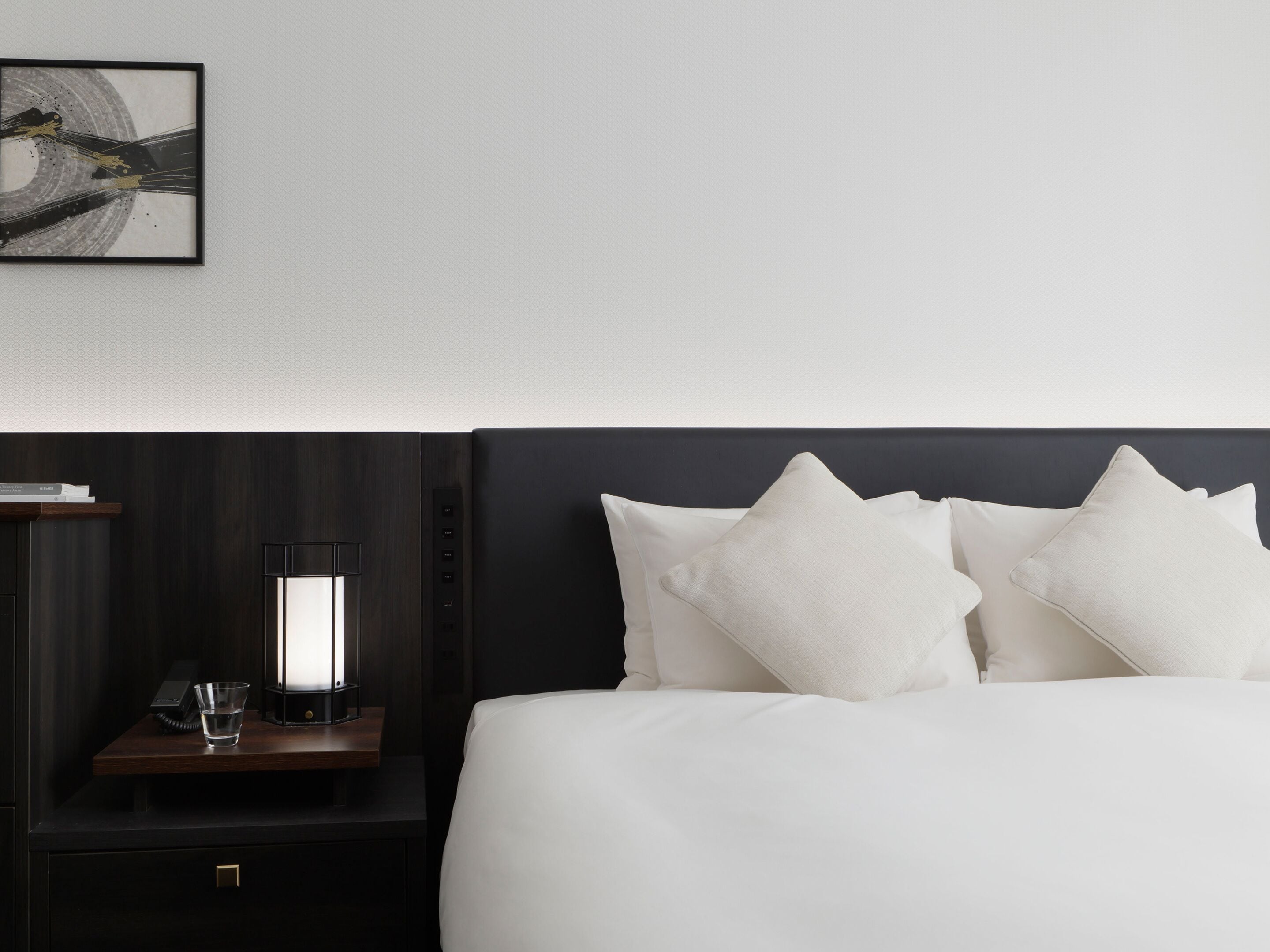 Un lit bien fait, avec du linge de lit blanc et plusieurs oreillers, une table de chevet avec une lampe, et un tableau encadré au mur.