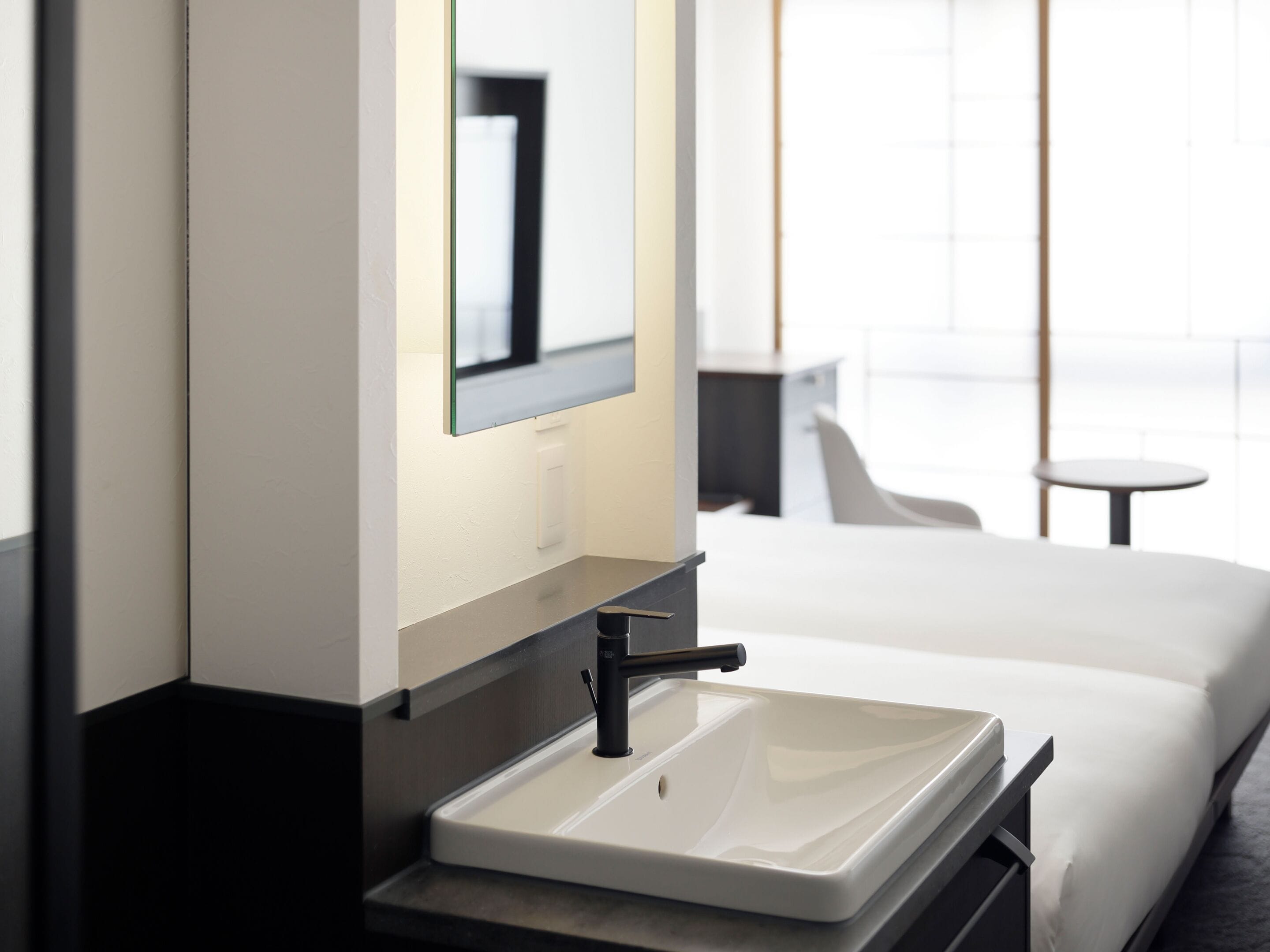 Un baño de hotel moderno con un lavamanos blanco, un espejo y una cama con ropa de cama blanca.