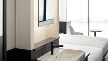 Un baño de hotel moderno con un lavamanos blanco, un espejo y una cama con ropa de cama blanca.