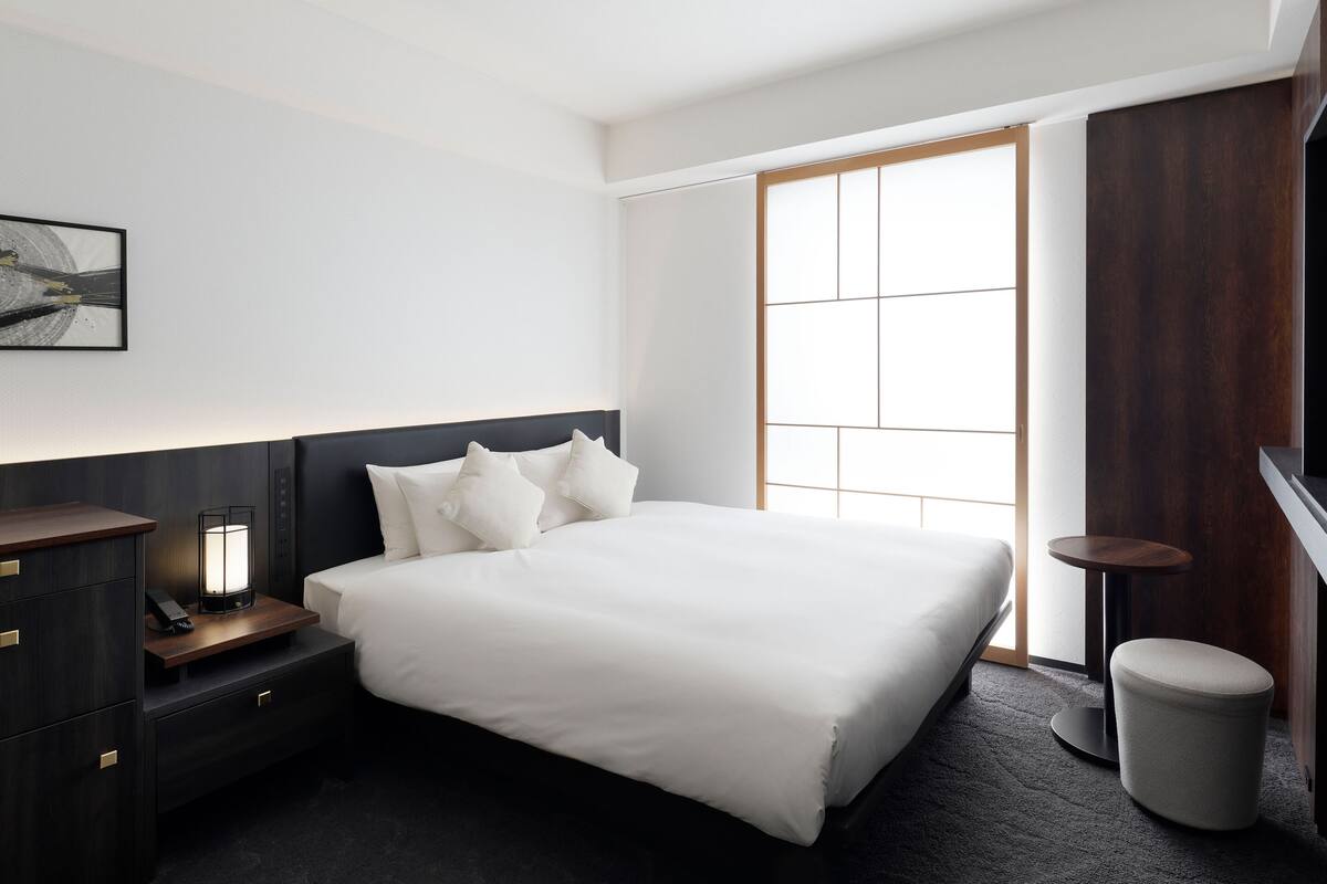 Et moderne hotelværelse med en stor seng, sengeborde, en lille skammel, en lampe og et vindue med udsigt.