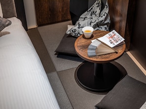 A neatly arranged hotel room with a bed, a round wooden table with a cup of tea, a fan, and a book.