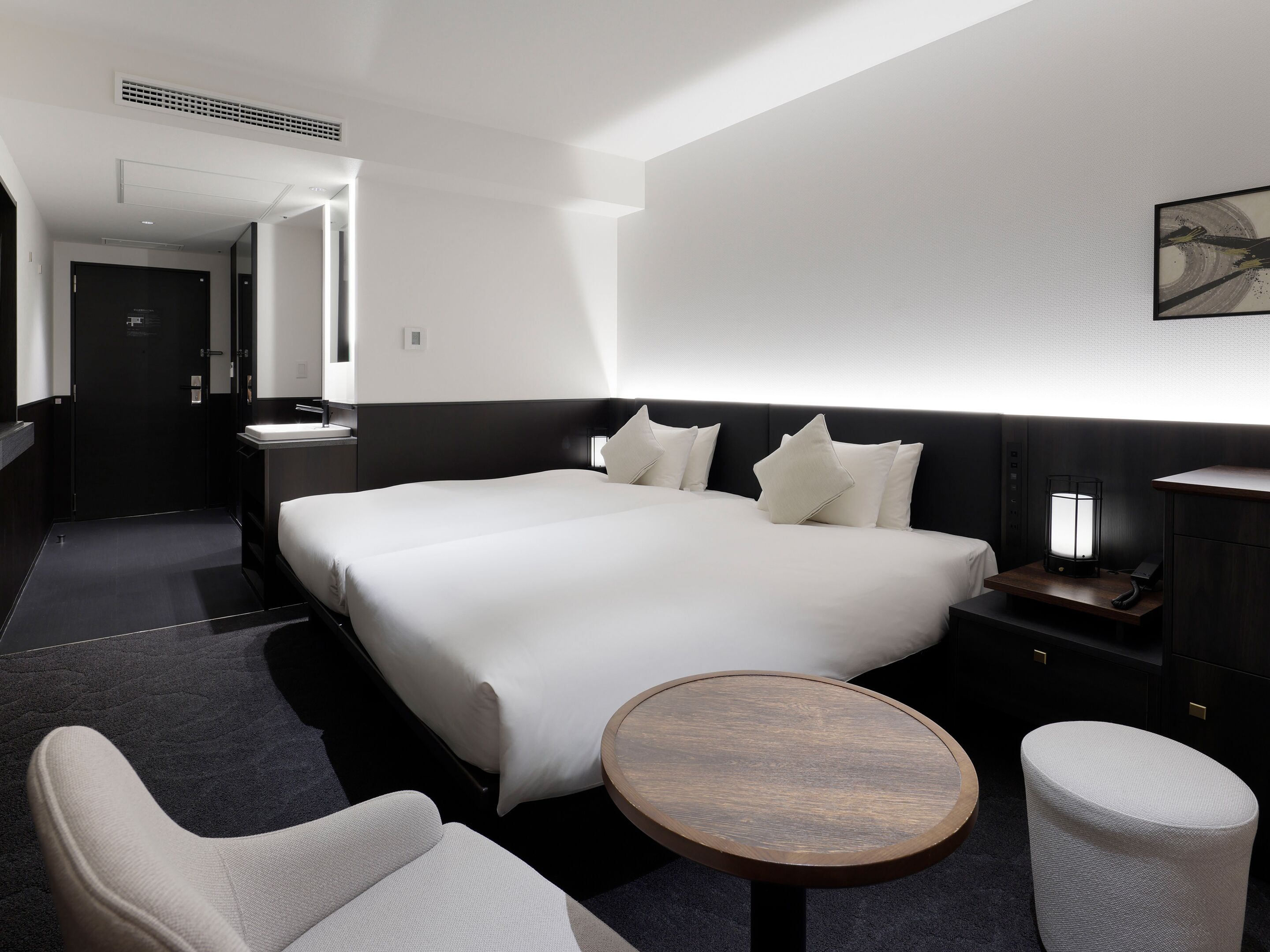 A hotel room with a large bed, bedside tables, a round wooden table, a chair, and a small stool.