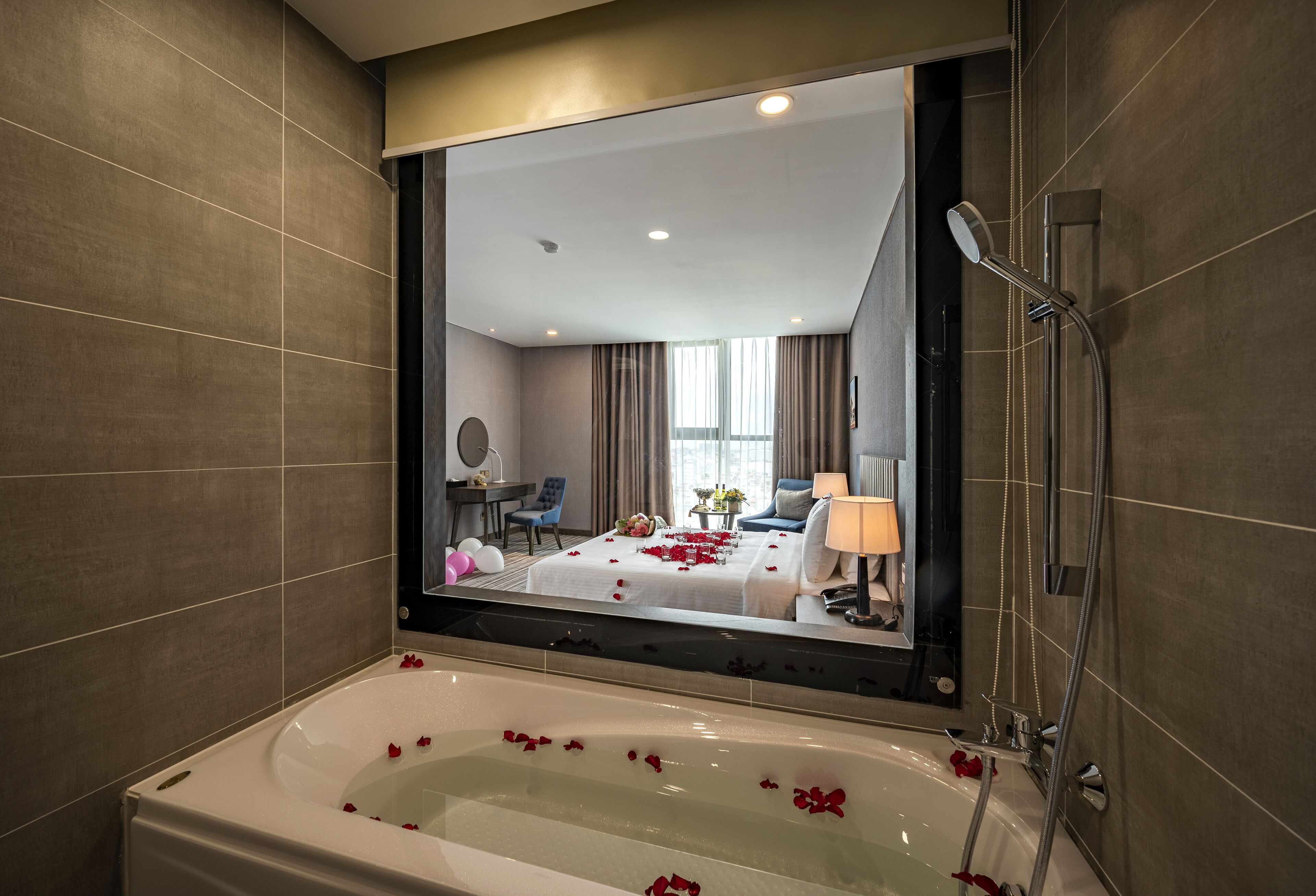 A modern bathroom with a large bathtub, a shower, and a mirror reflecting a bedroom with a bed, a desk, and a chair.