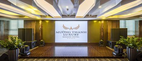 A hotel lobby with a stage, potted plants, and a large sign reading "MUONG THANH LUXURY SAIGON HOTEL".