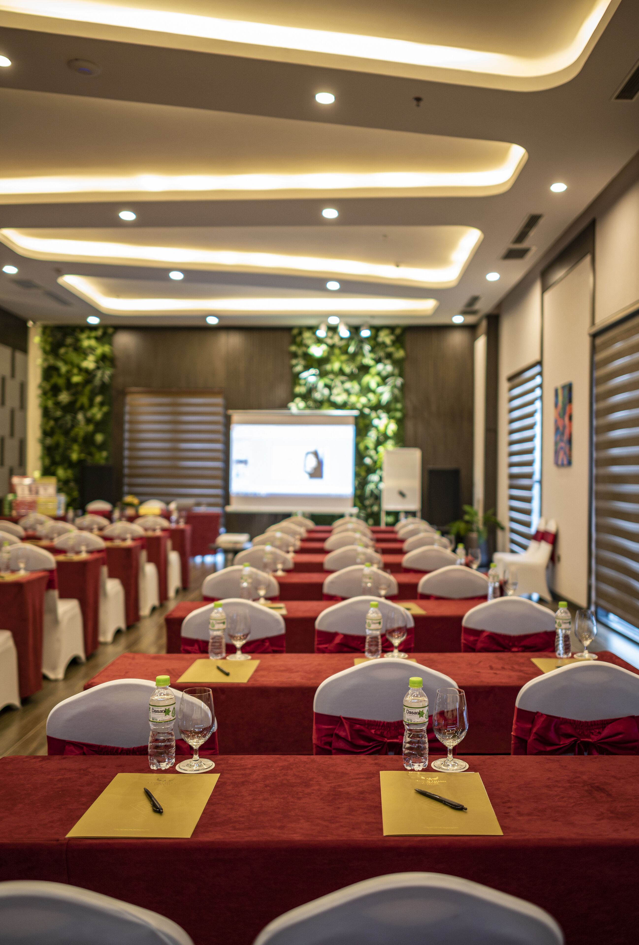 A conference room with rows of tables, each with a red tablecloth, white chairs, and water bottles. A projector screen is visible in the background.