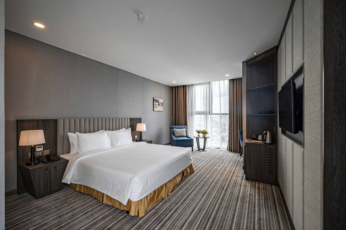 A hotel room with a large bed, a desk with a lamp, a chair, a TV, and a window with curtains.