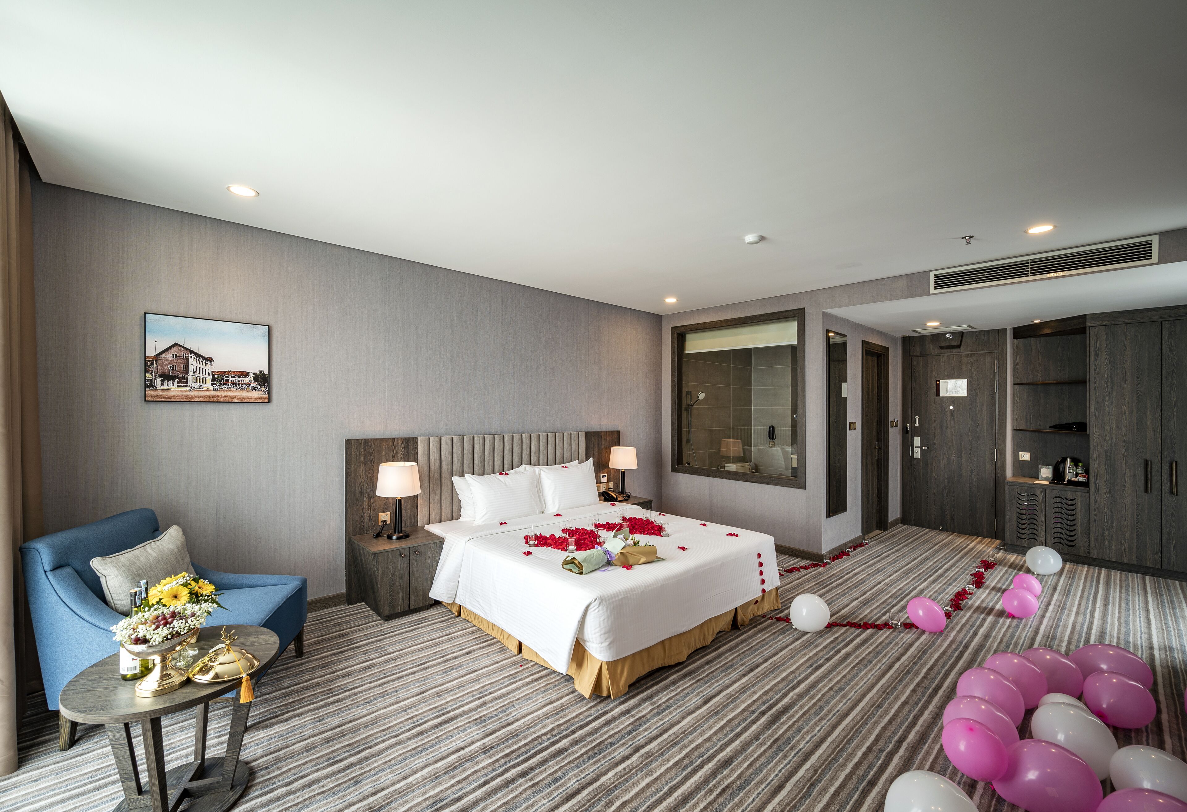A hotel room with a bed, a blue armchair, a small table with a vase of flowers, and a large window.
