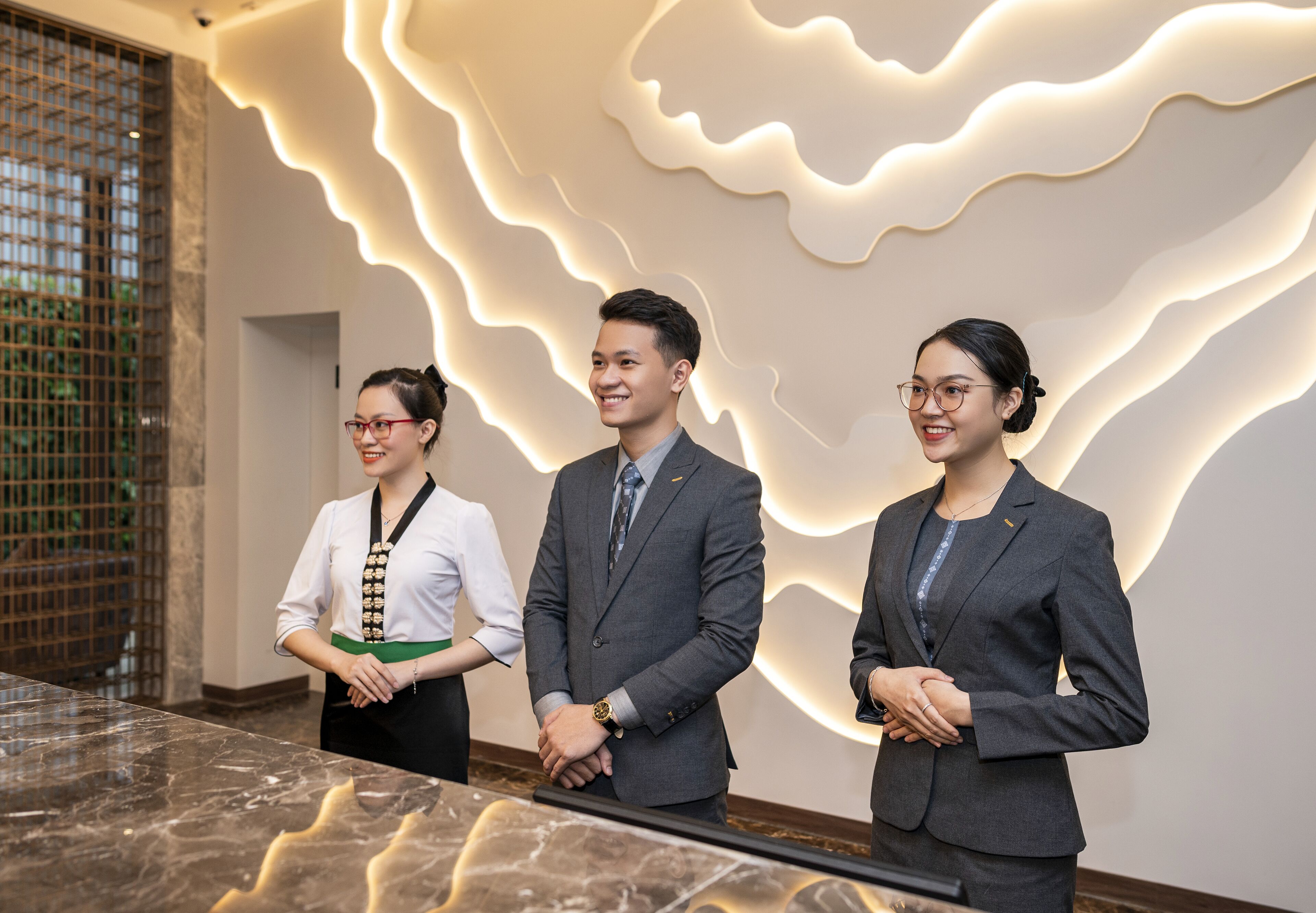 Three individuals standing in a lobby with a modern, wavy light design on the wall and a marble floor.