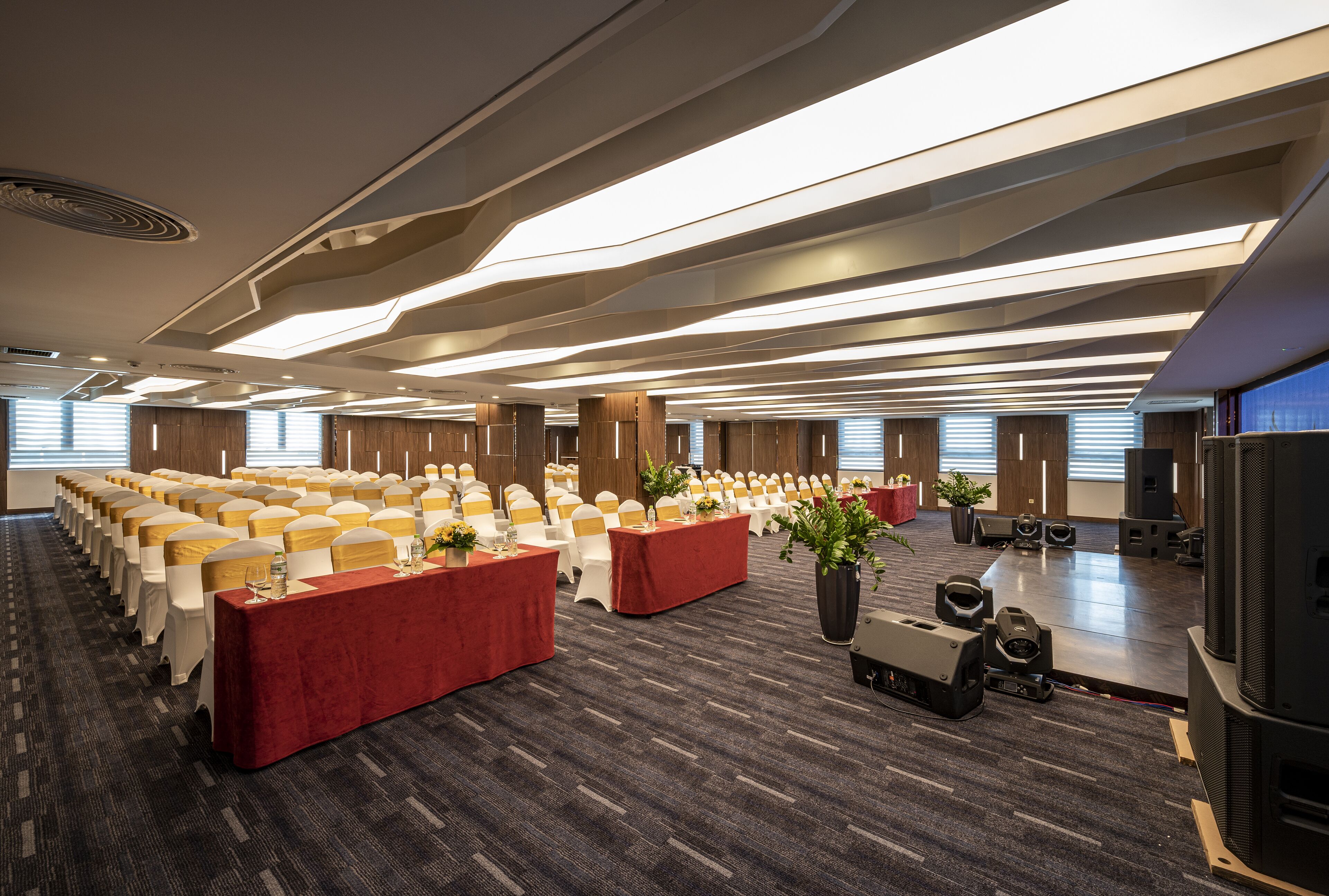 Un salón de conferencias con filas de sillas blancas, mesas rojas y un escenario con equipo de audio.