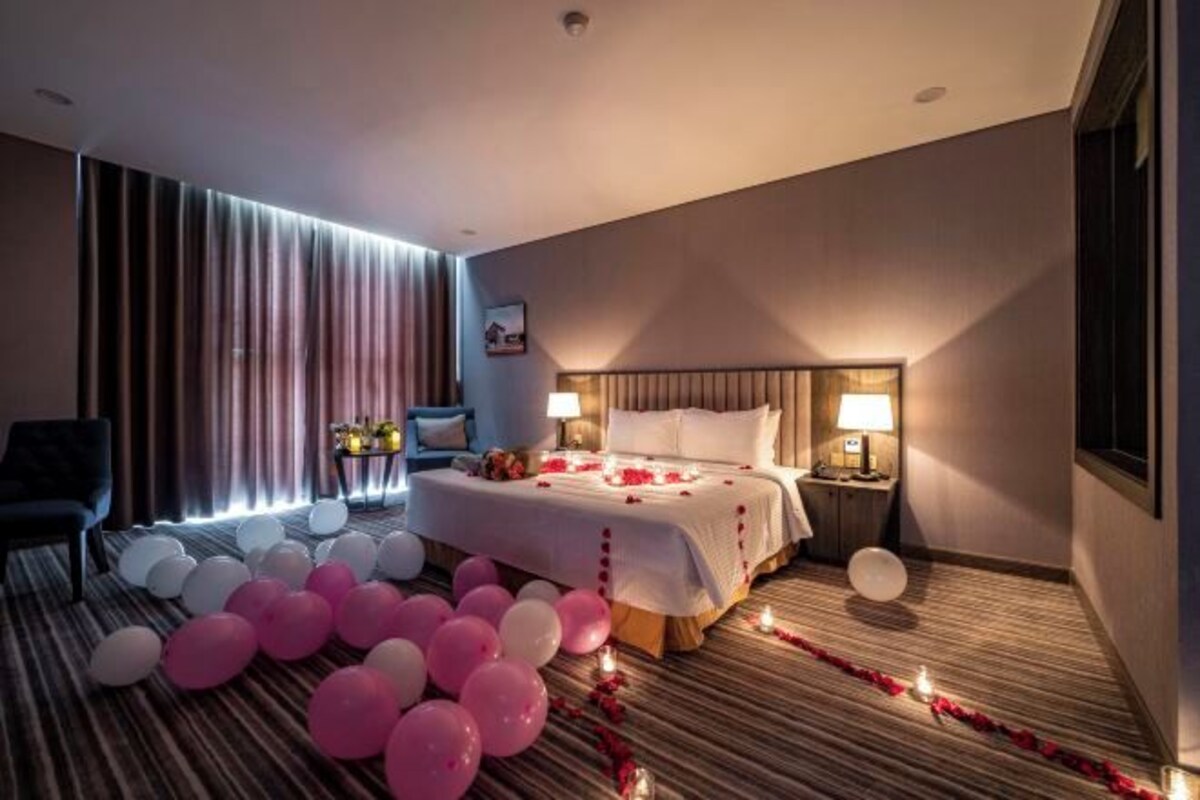 A hotel room with a large bed, a bedside table with a lamp, a chair, and a small table with a vase of flowers.