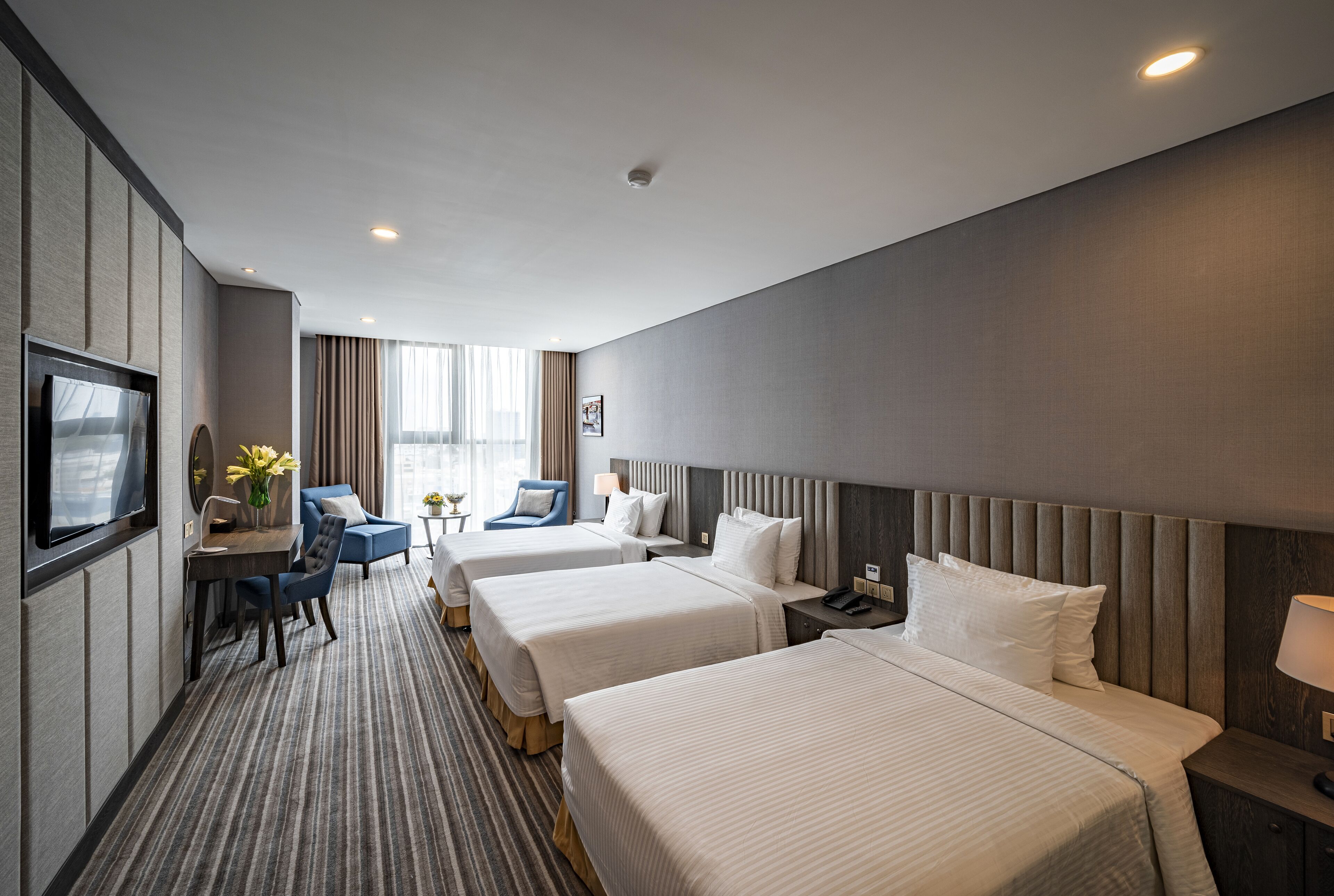 A hotel room with three beds, a desk with a chair, a TV, and a window with curtains.