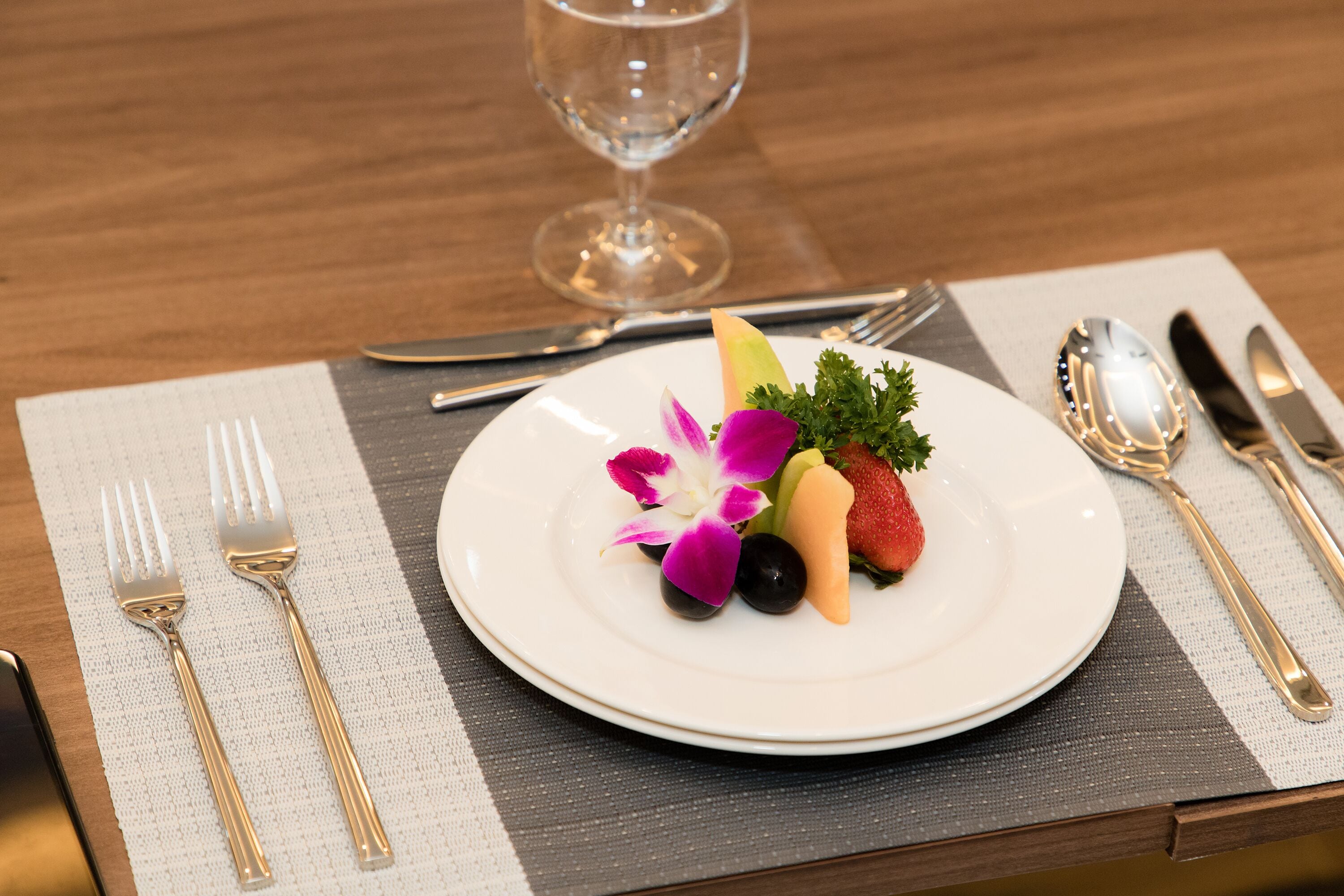A neatly arranged dining table with a white plate of fruit, a glass of water, and a wine glass.