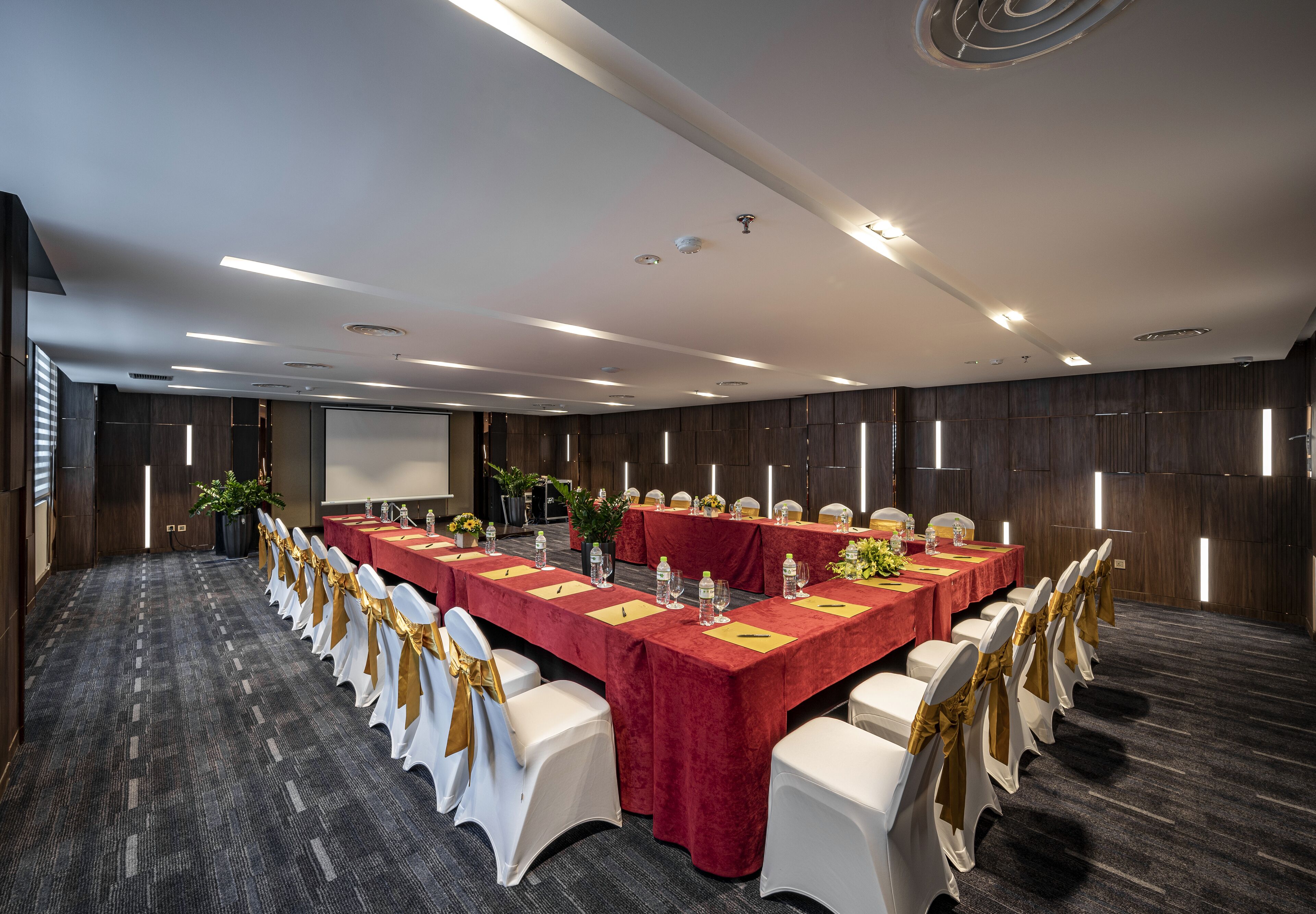 Une salle de conférence avec une longue table, des chaises blanches ornées de rubans dorés et une nappe rouge.
