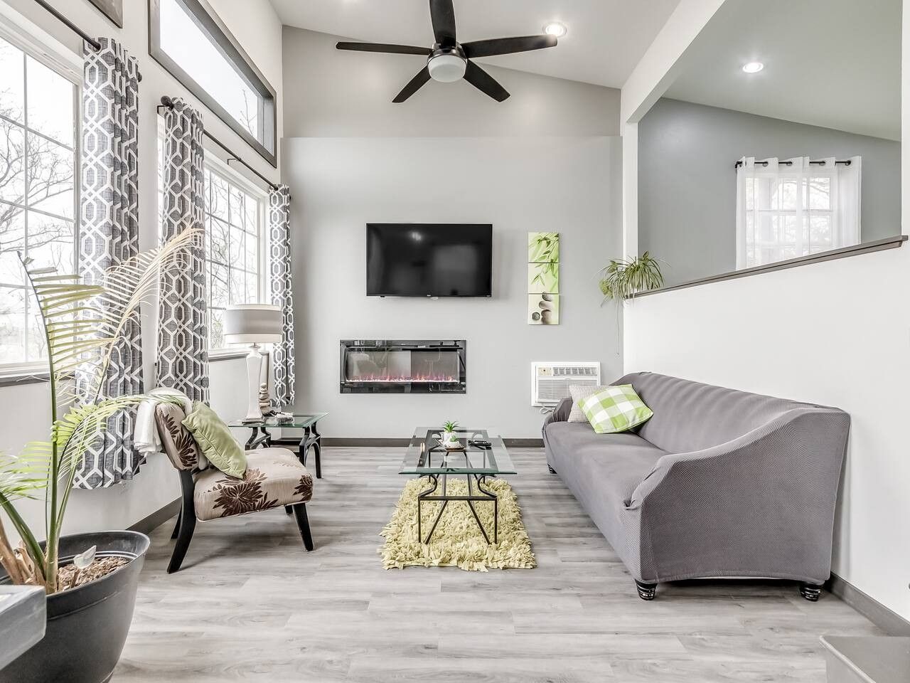 A modern living room with a grey sofa, a patterned armchair, a flat-screen TV, a fireplace, and a ceiling fan.
