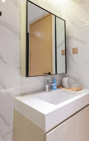 Een badkamer met een witte marmeren wand, een houten deur, een witte wasbak, een spiegel en een blauwe handzeepfles.