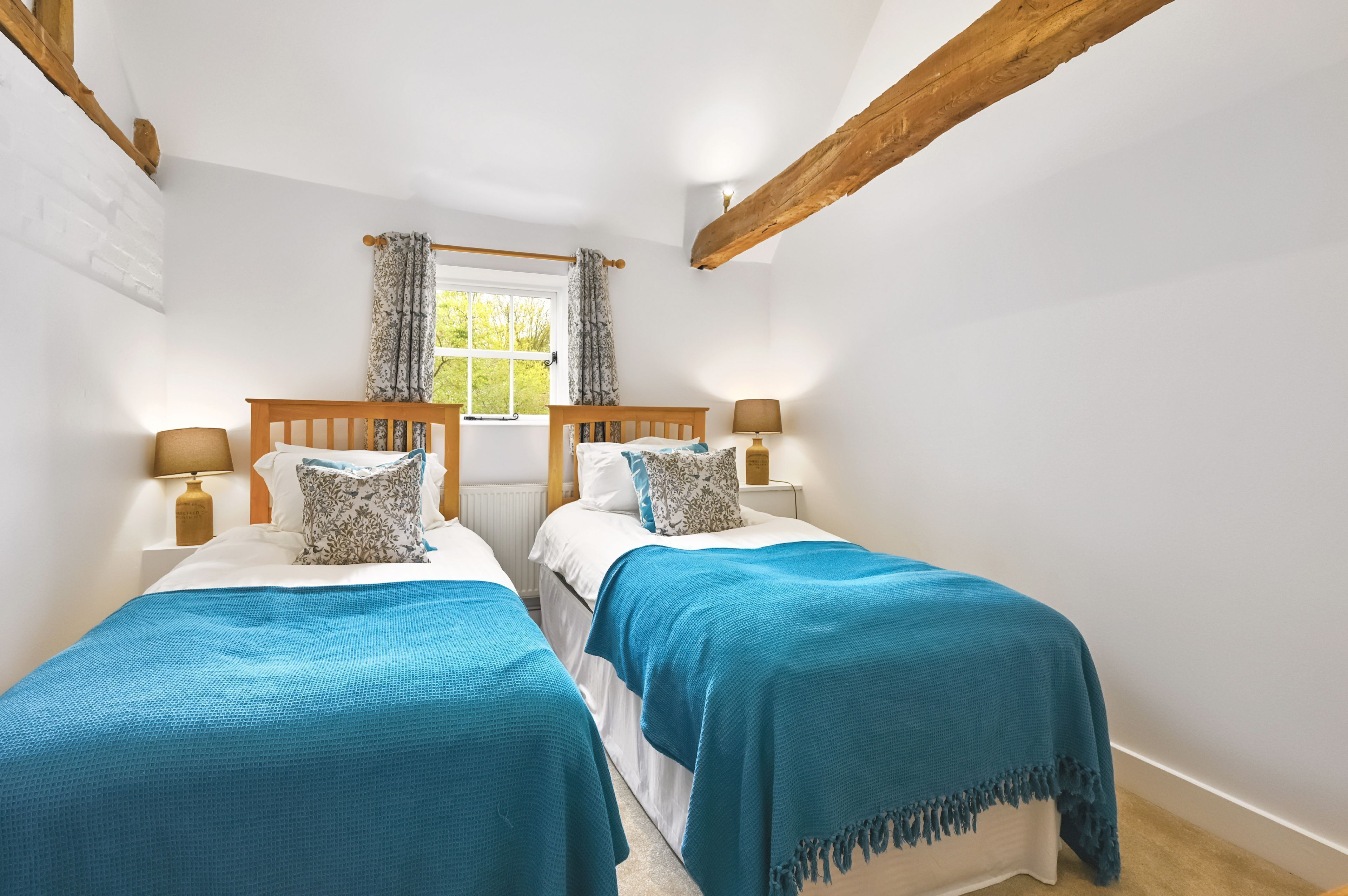 A room with two beds, wooden beams, a window with curtains, and a white brick wall.