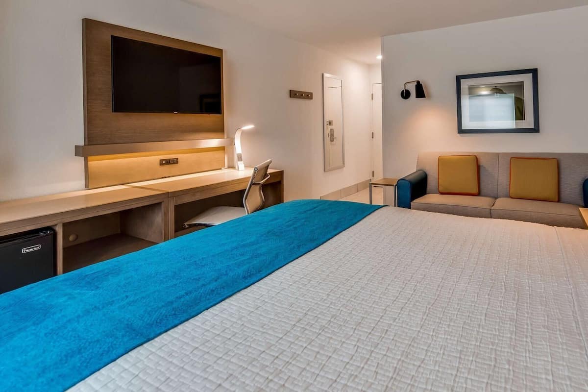 Habitación de hotel con cama, escritorio, silla, sofá, televisión y lámpara de pared.