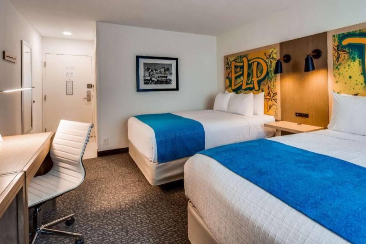 Habitación de hotel con dos camas, un escritorio, una silla y una pared con una imagen de un paisaje urbano y un gran cartel que dice 'ELP'.