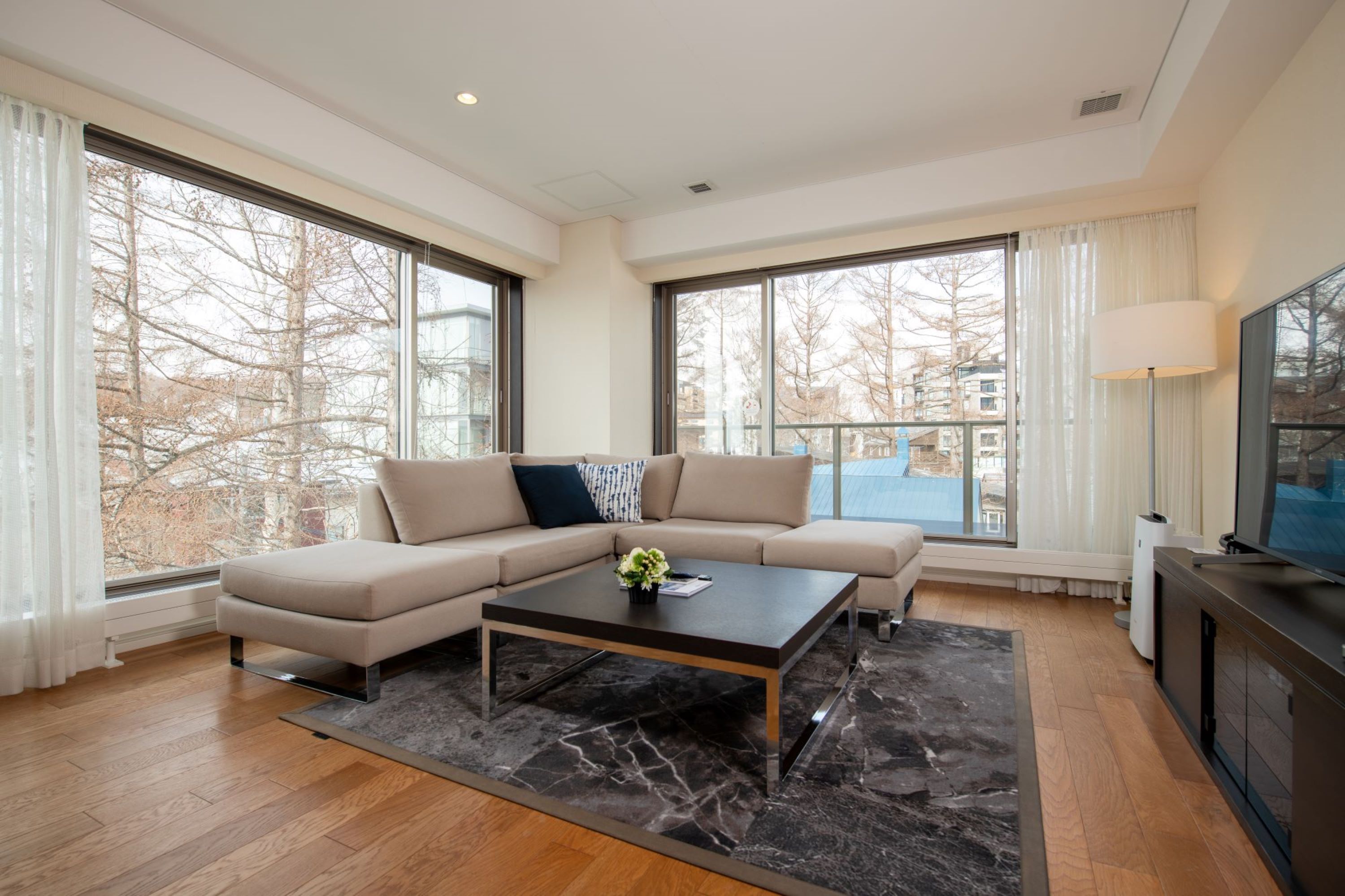 Un salon moderne avec un canapé d’angle, une table basse et de grandes fenêtres donnant sur l’extérieur.