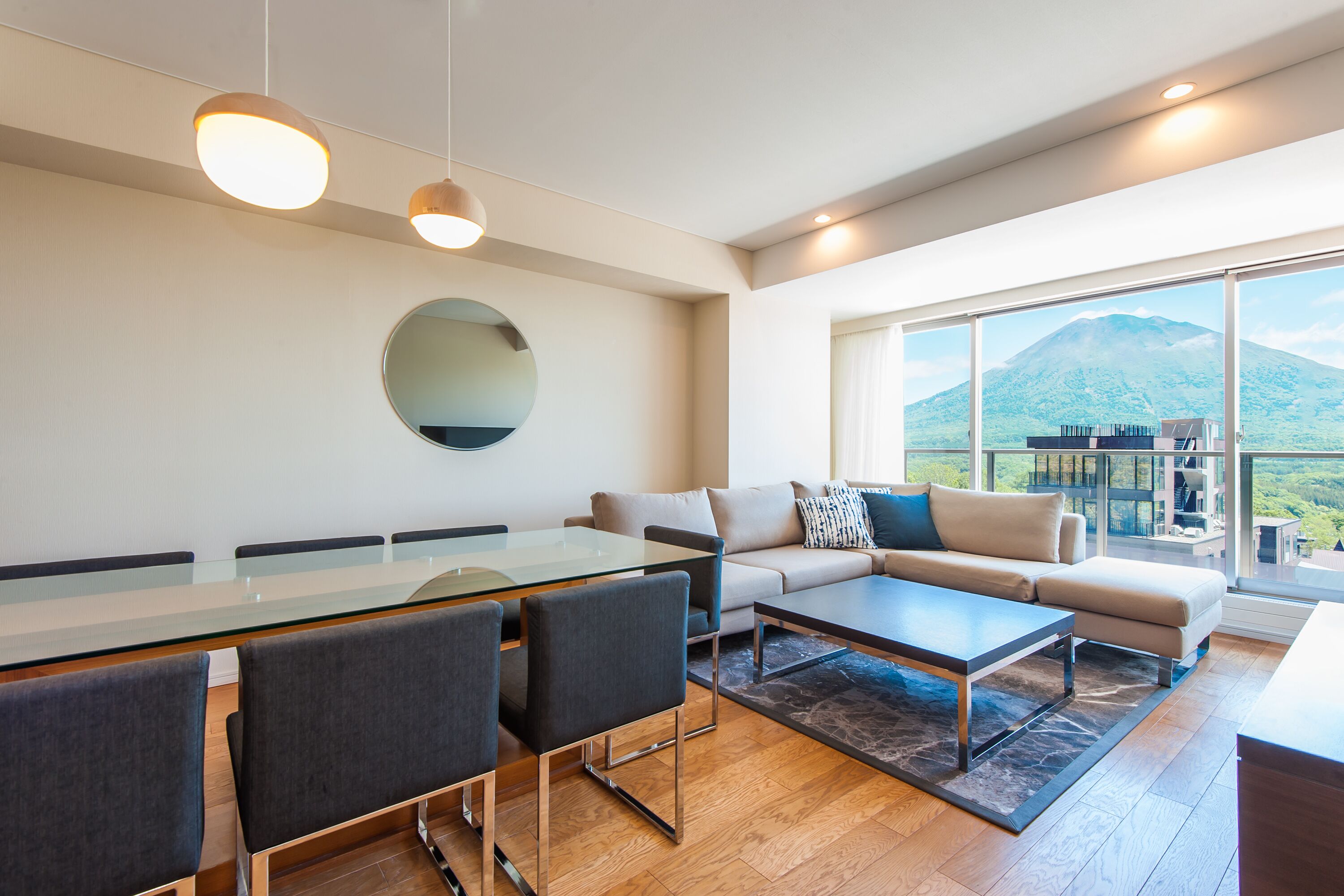 A modern living room with a glass dining table, grey sofas, and a large window offering a mountain view.