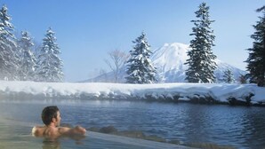 Una persona en un manantial termal rodeada de árboles cubiertos de nieve y una montaña al fondo.