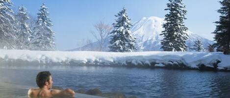 A person in a hot spring surrounded by snow-covered trees and a mountain in the background.