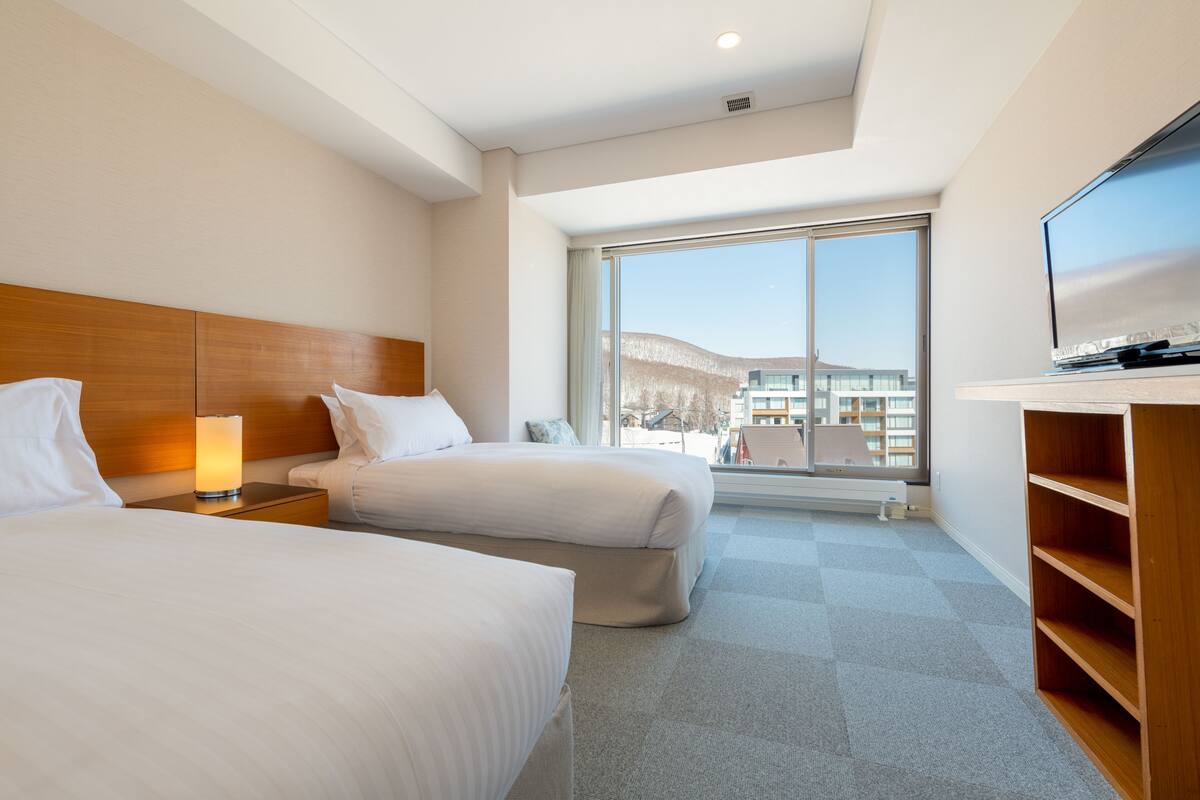 Habitación de hotel con dos camas, televisión, ventana grande y vista a edificios.