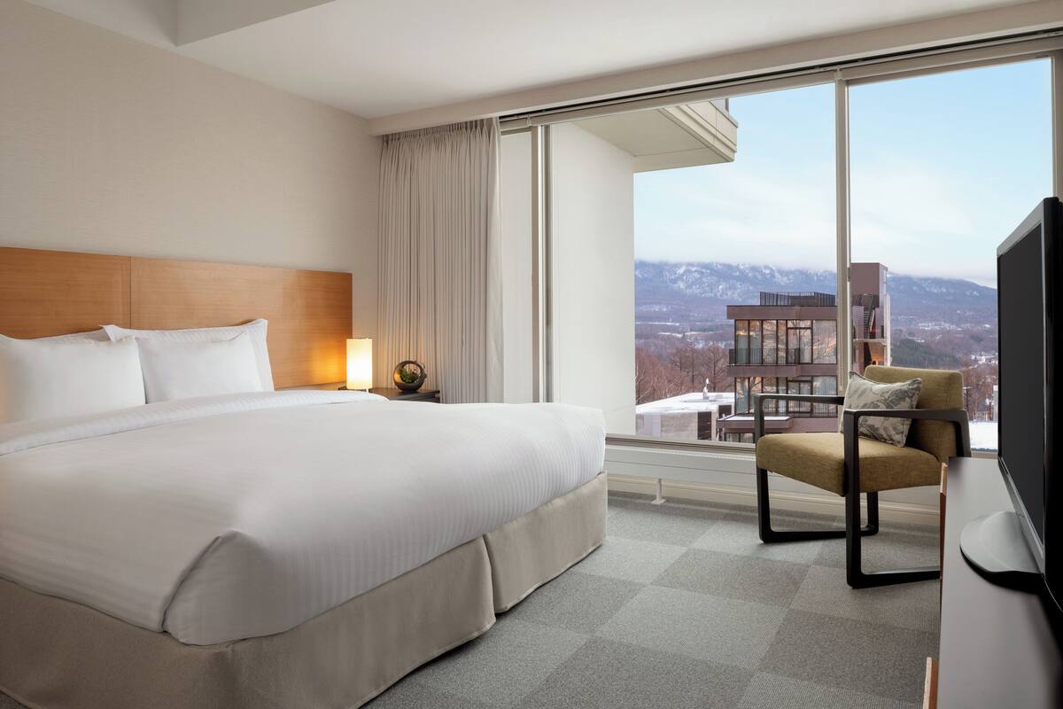 Habitación de hotel con una cama grande, una silla, un televisor y vistas a un paisaje urbano a través de amplias ventanas.