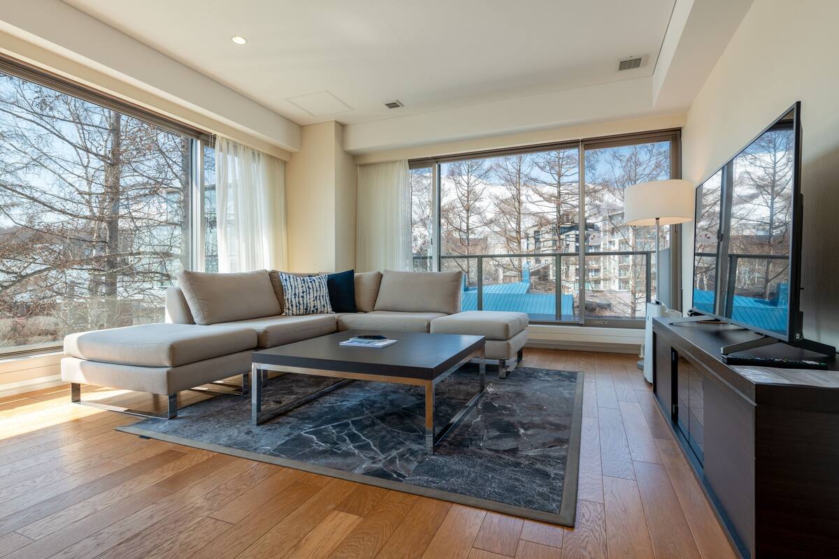 Una sala de estar moderna con un sofá seccional, una mesa de centro y un televisor sobre un soporte. Amplias ventanas ofrecen vistas al exterior.