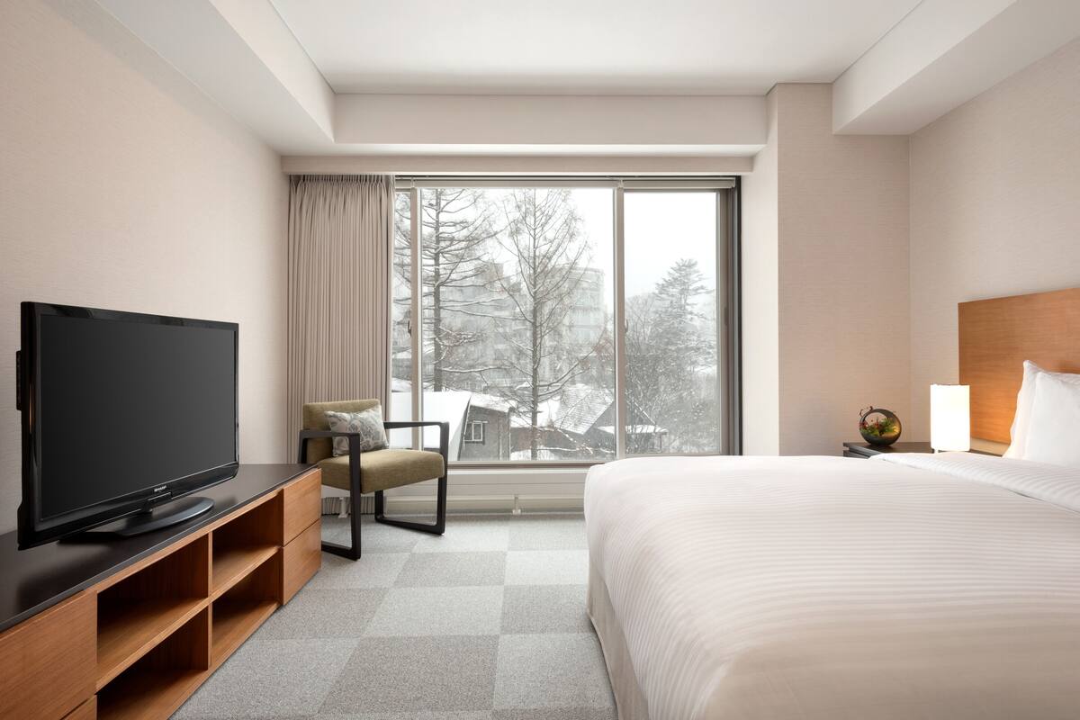 Habitación de hotel con cama, televisor sobre un mueble de madera, una silla, mesita de noche y un ventanal con vistas a edificios y árboles.