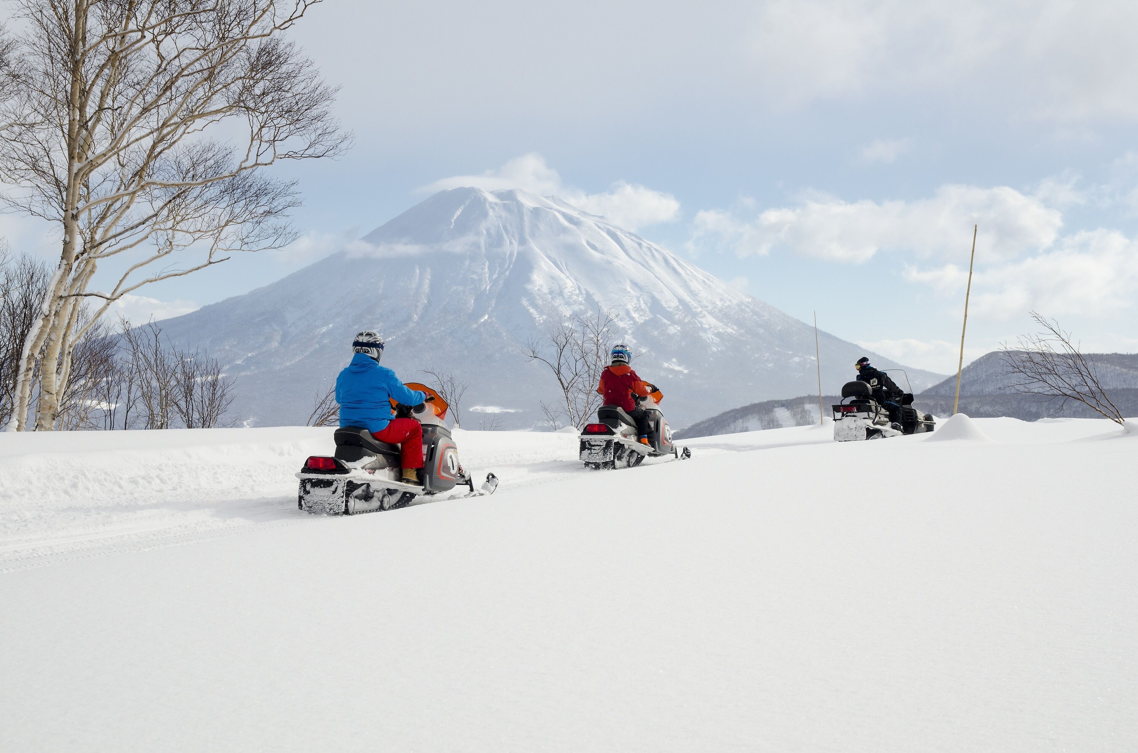 Three people on snowmobiles on a snowy path with a mountain in the background.