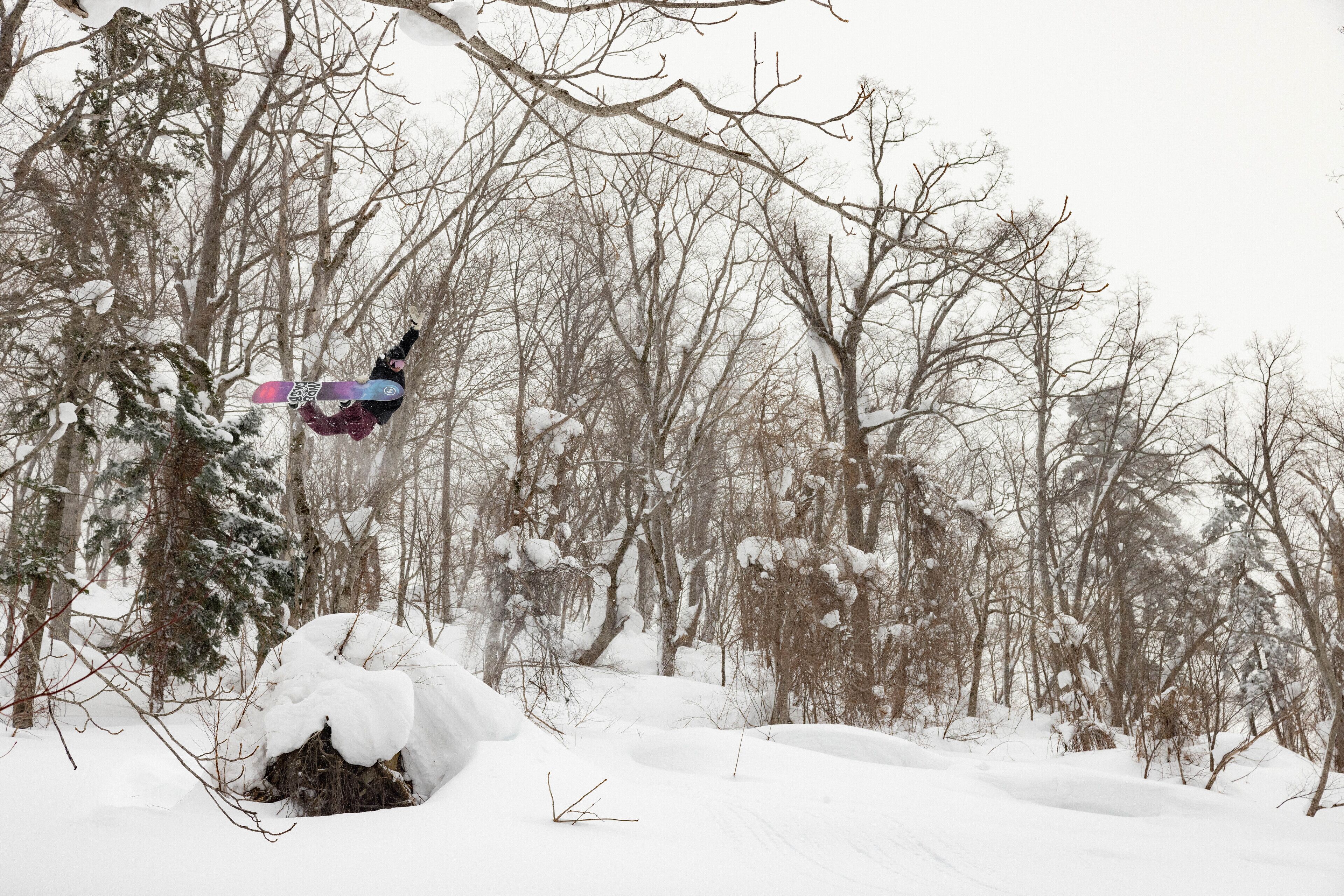 Eine verschneite Waldszene, ein Snowboarder in der Luft.