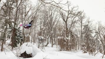 Eine verschneite Waldszene, ein Snowboarder in der Luft.