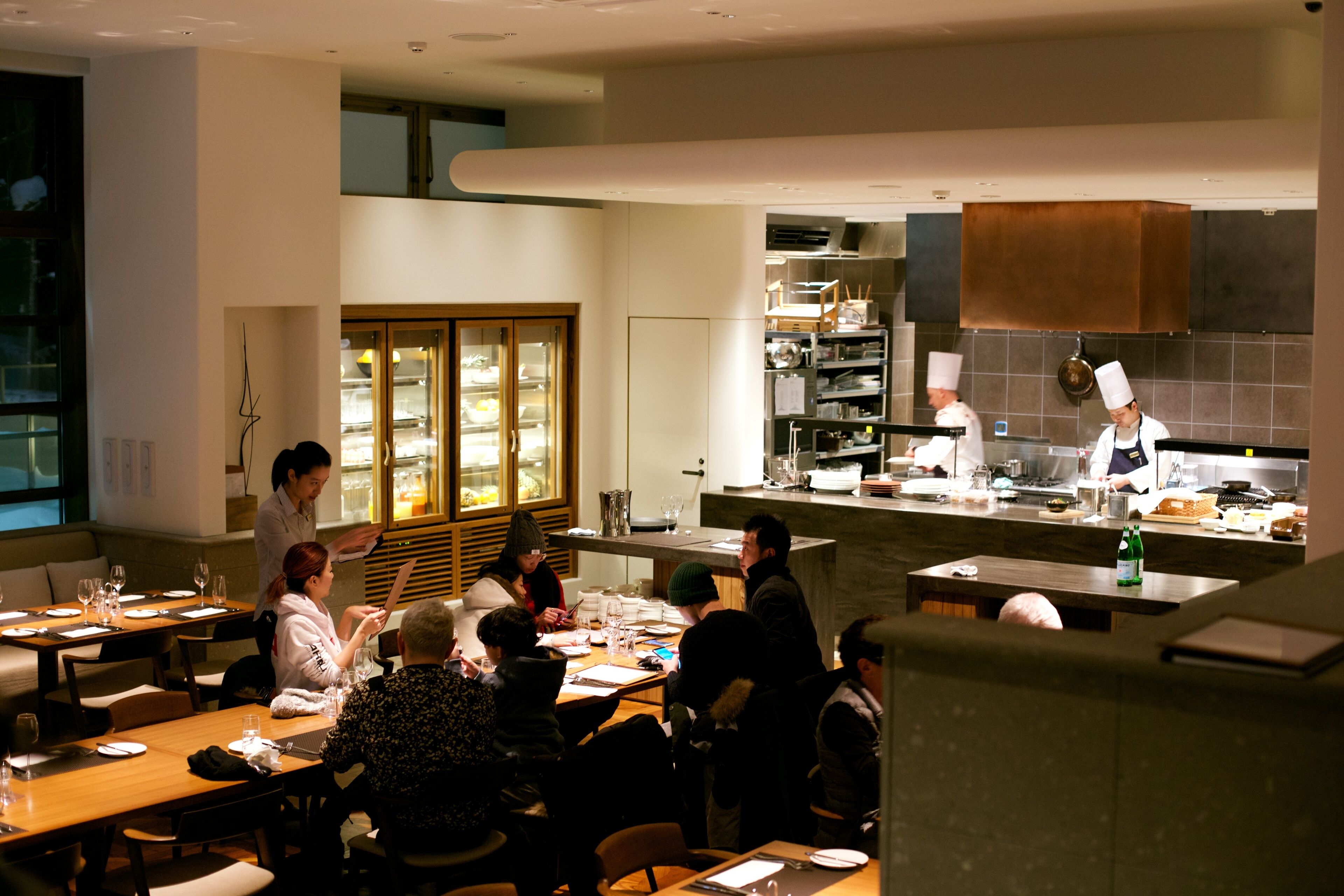 A modern restaurant with a chef preparing food at the counter, diners seated at tables, and a view of the kitchen area.
