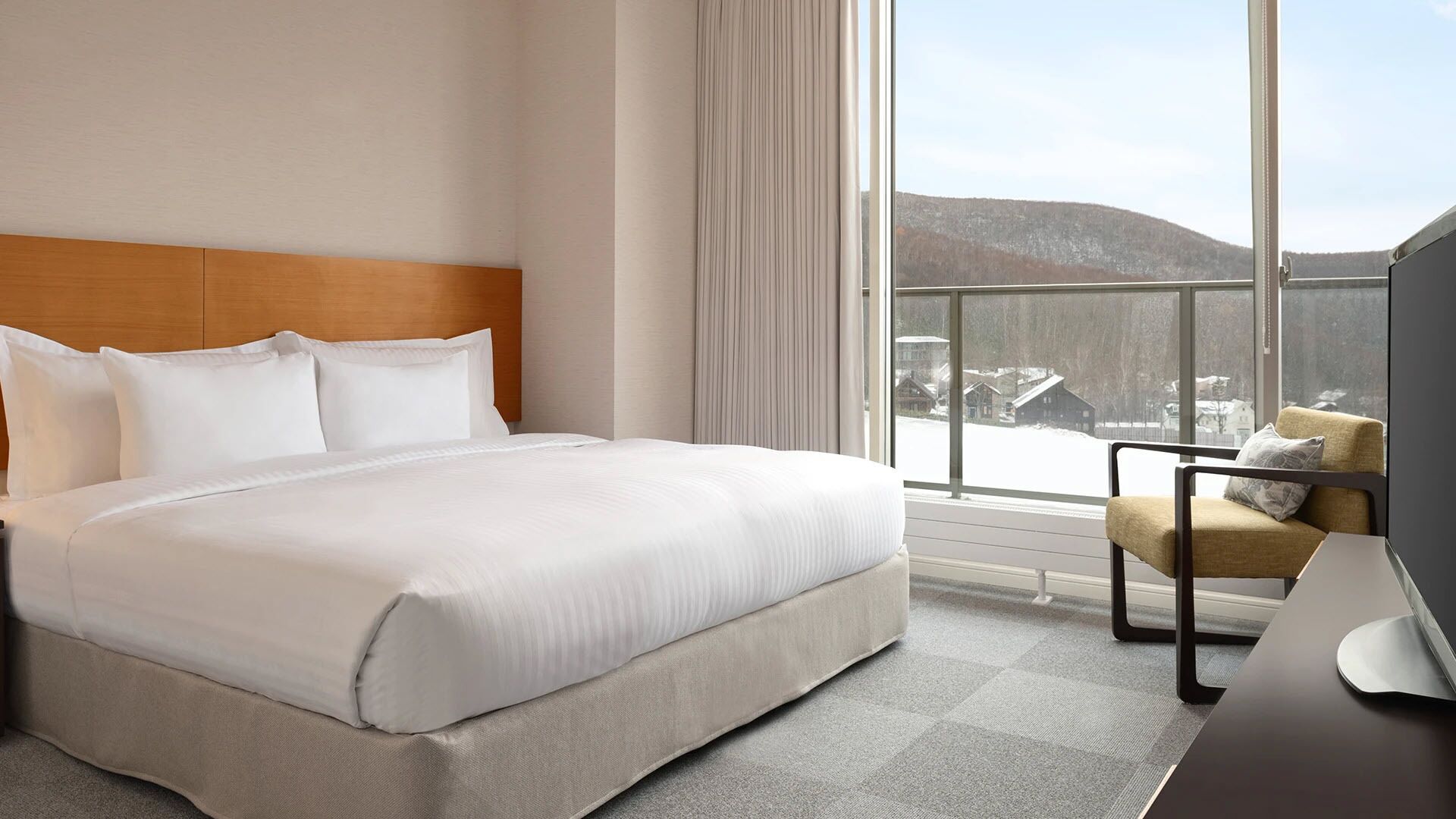 A hotel room with a large bed, a chair, a desk, and a window with a view of snow-covered houses and hills.