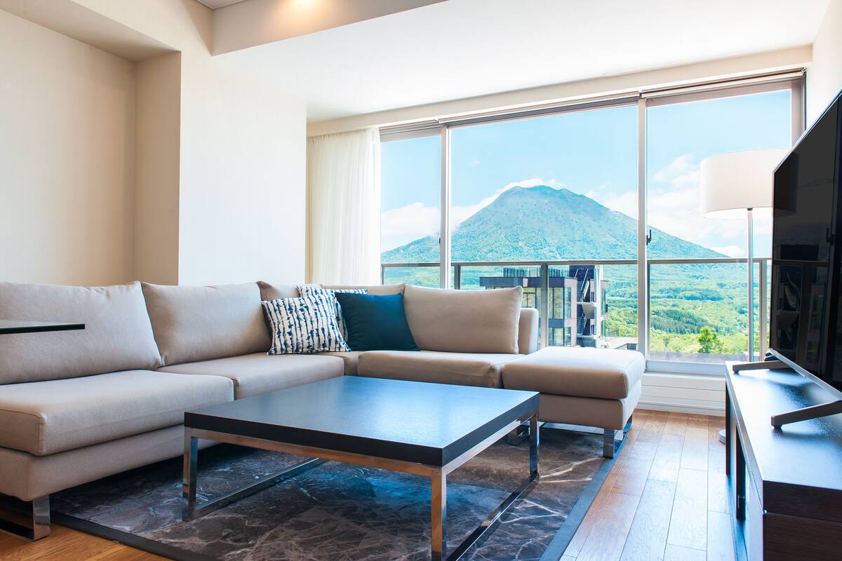 Una sala de estar moderna con un sofá, una mesa de centro y un televisor. Se ve una ventana grande con vistas a una montaña.